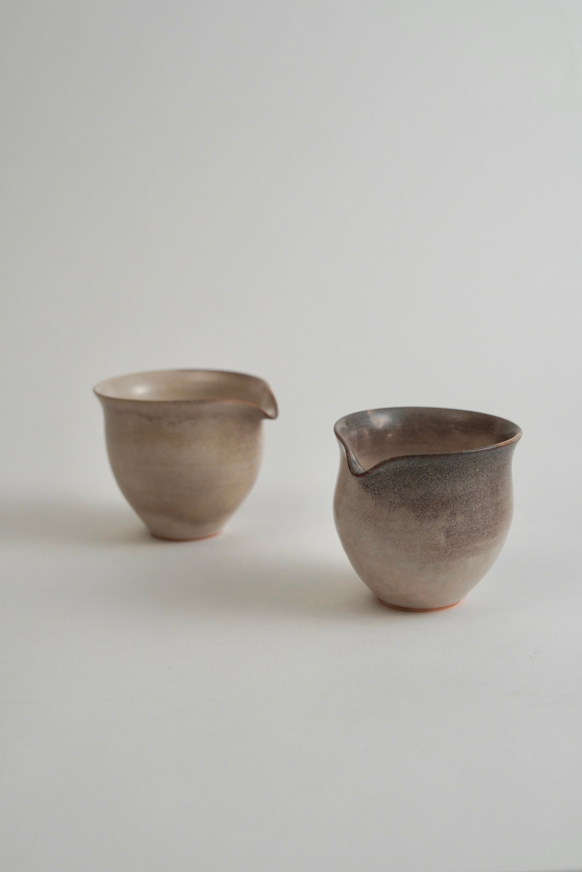 Two handmade ceramic pouring cups with a matte, earthy finish, displayed on a neutral light gray background.