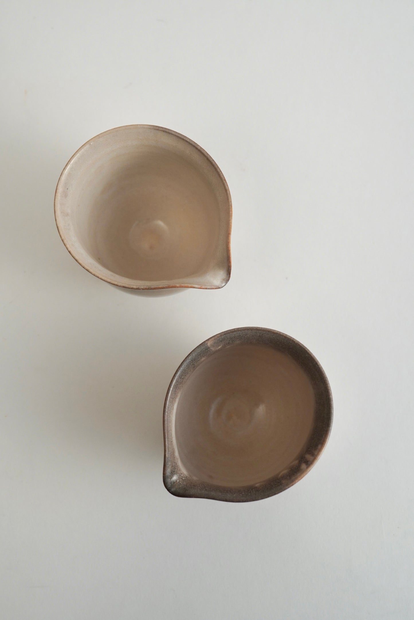 Two small ceramic bowls with spouts, shown from above on a plain white background. One bowl has a light beige interior and exterior, while the other has a darker brown interior and exterior.