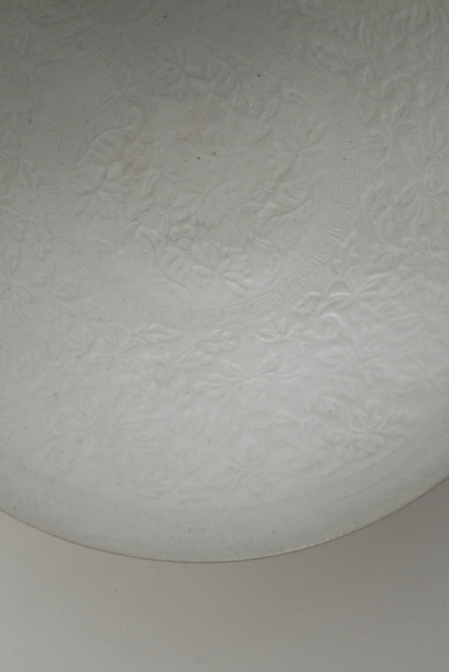 A close-up of a white textured glass or acrylic surface with a subtle embossed floral pattern, showing a curved edge and a neutral light background.