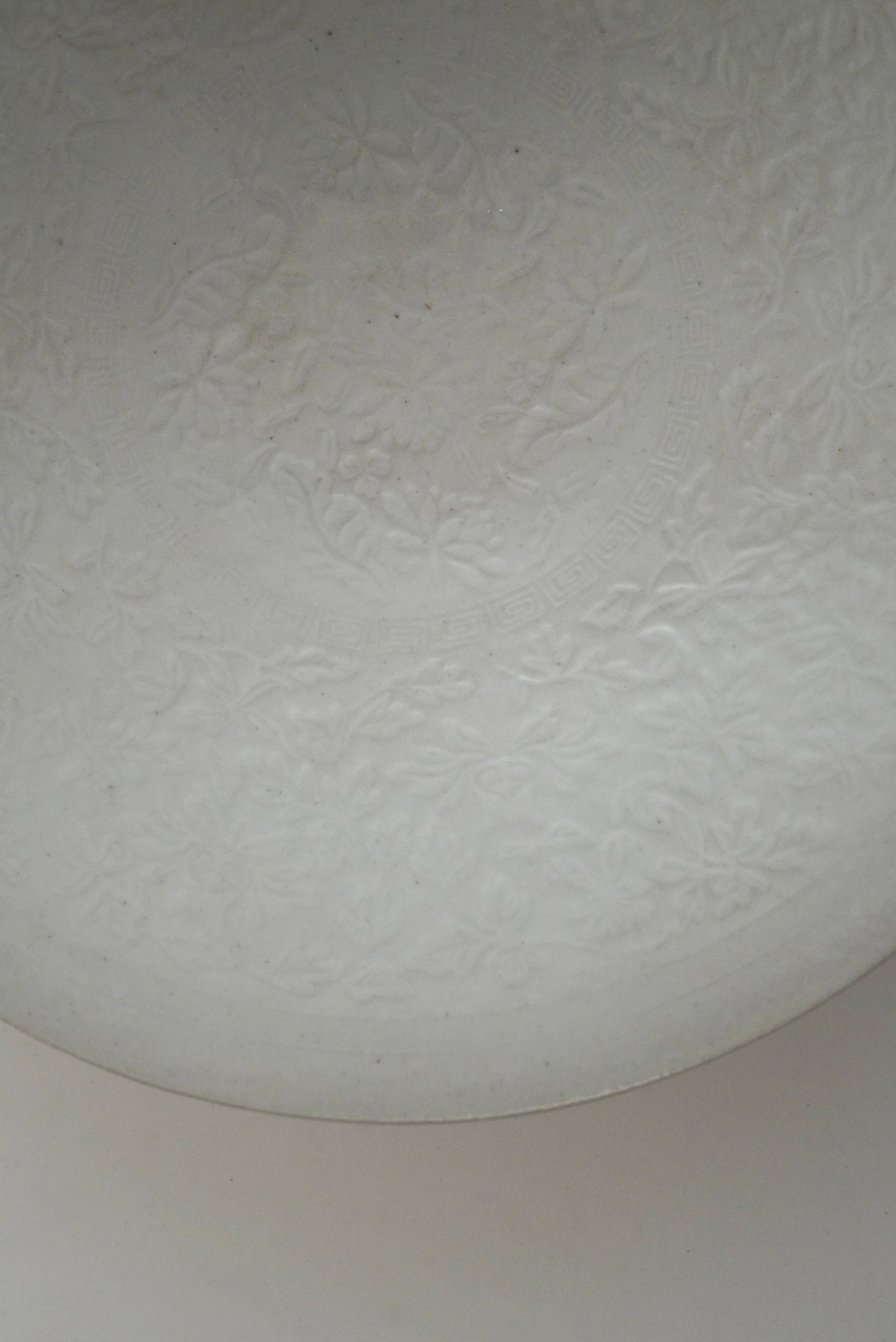 A close-up of a white textured glass or acrylic surface with a subtle embossed floral pattern, showing a curved edge and a neutral light background.