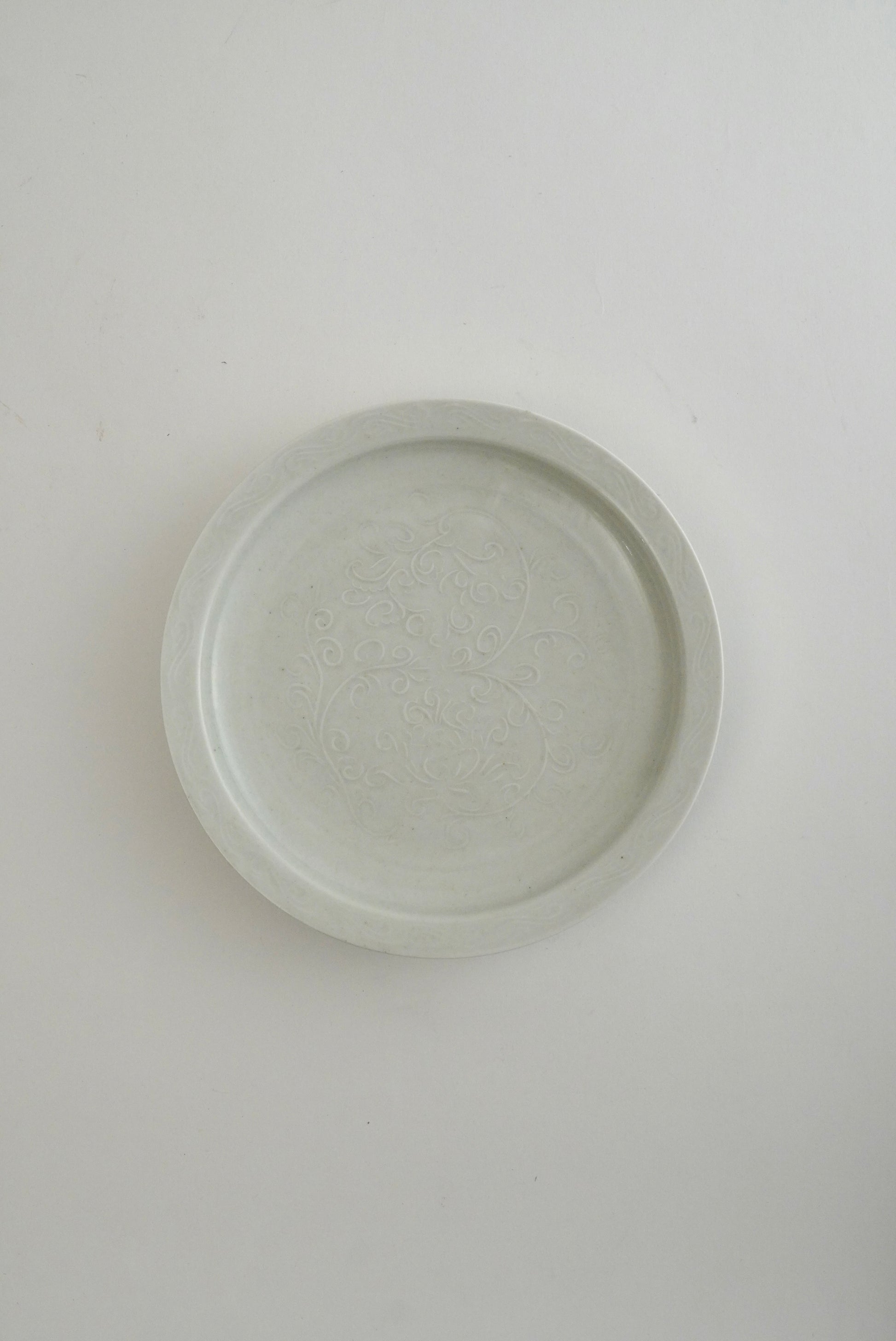 A round, shallow ceramic plate or dish with a subtle embossed floral pattern, shown from a top-down perspective on a plain white background.