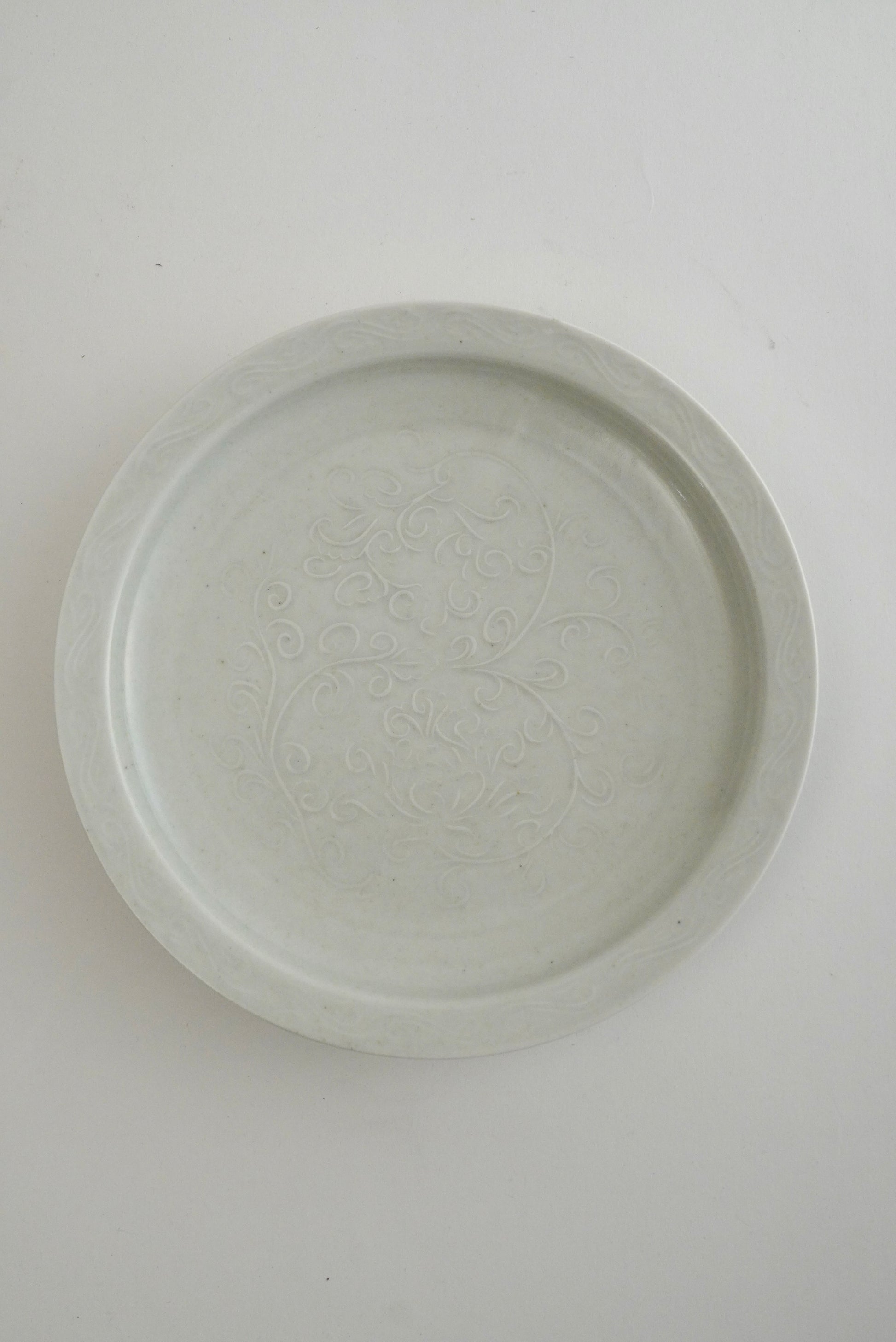 A round, shallow ceramic plate or dish with a subtle embossed floral pattern, shown from a top-down perspective on a plain white background.