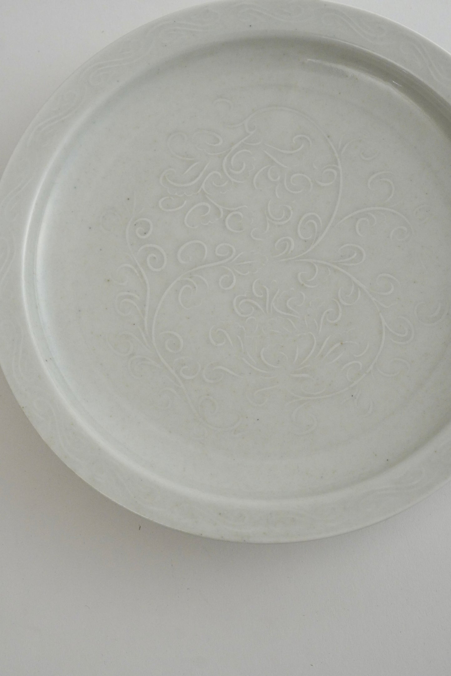 A round white ceramic plate with a subtle embossed floral pattern in the center, photographed from directly above on a plain white background.