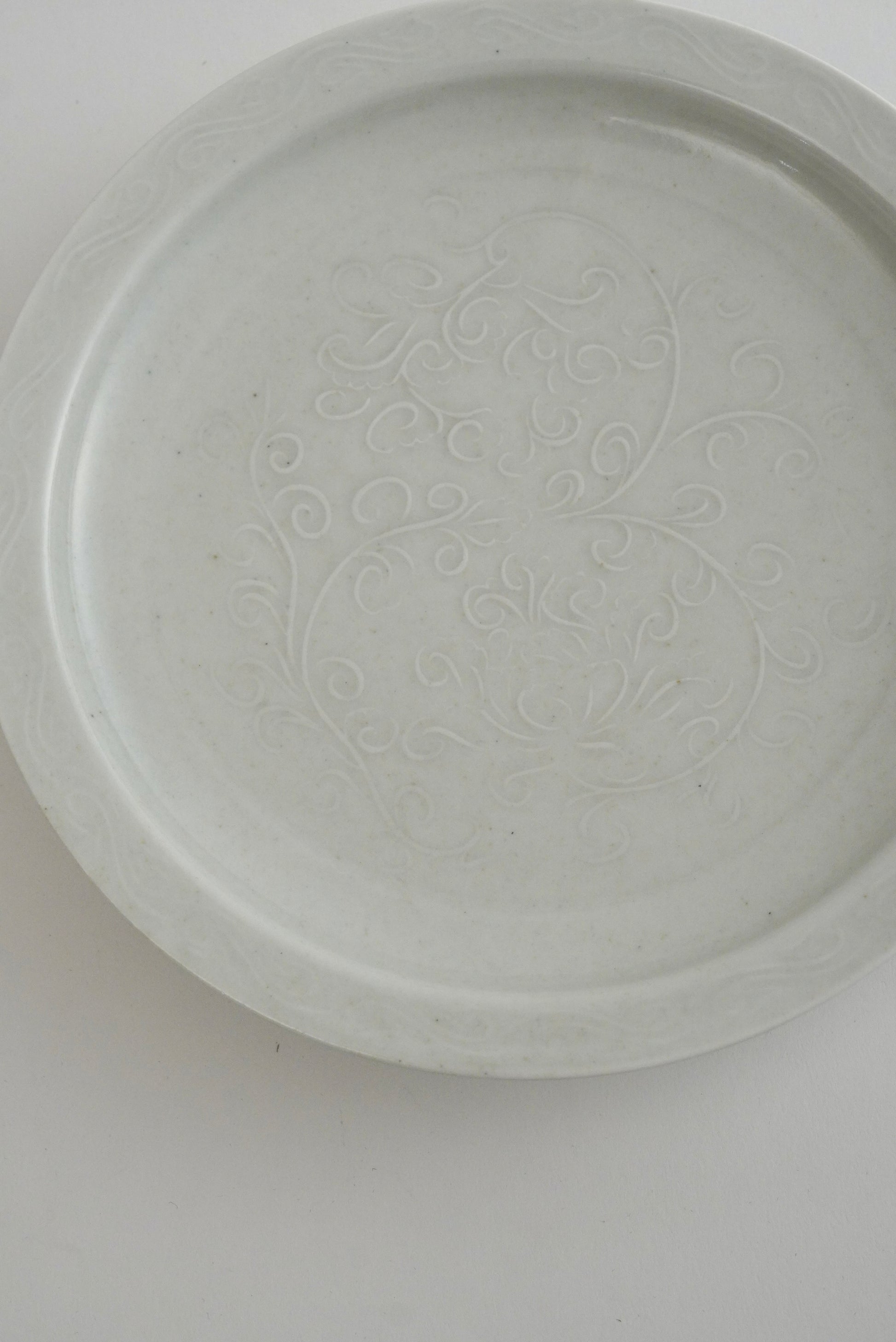 A round white ceramic plate with a subtle embossed floral pattern in the center, photographed from directly above on a plain white background.