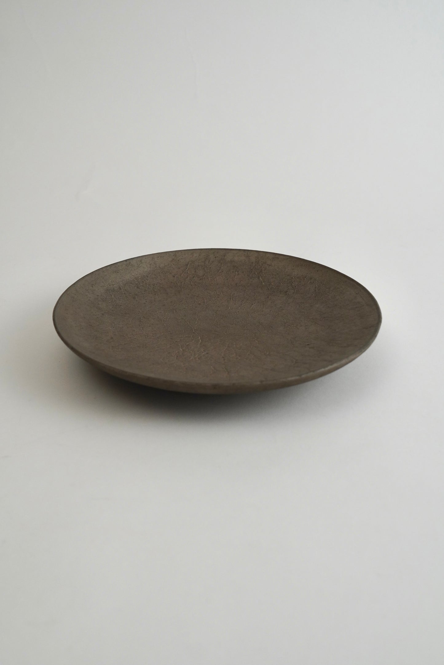 A round, shallow dark brown ceramic plate with a matte, textured finish, photographed on a neutral light gray background.