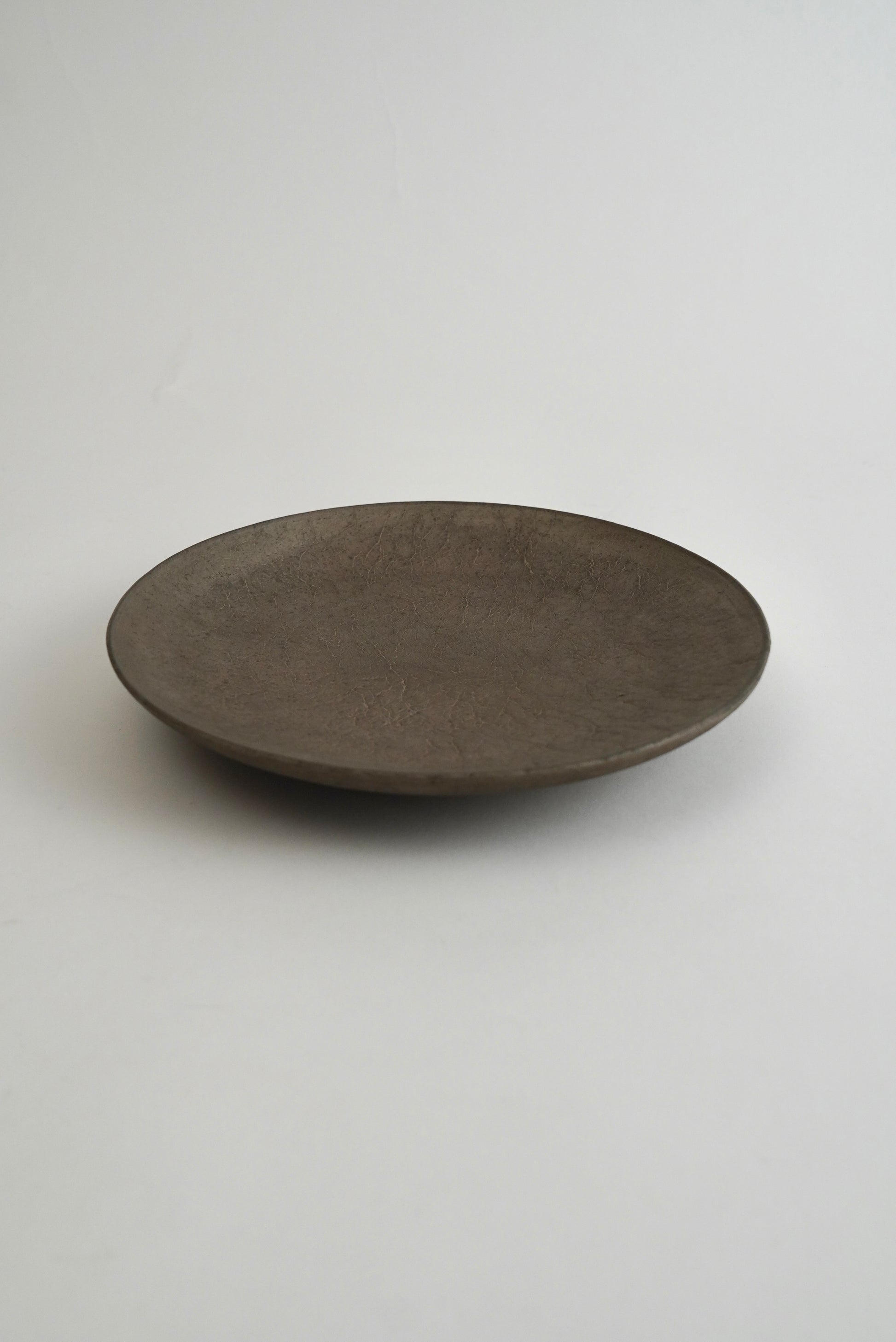 A round, shallow dark brown ceramic plate with a matte, textured finish, photographed on a neutral light gray background.