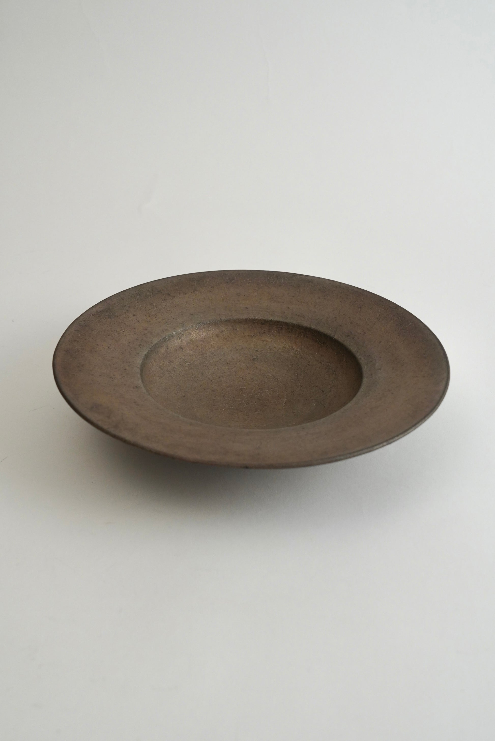 A round, shallow ceramic bowl with a wide rim, featuring a matte, dark brown textured glaze, set against a plain light gray background.
