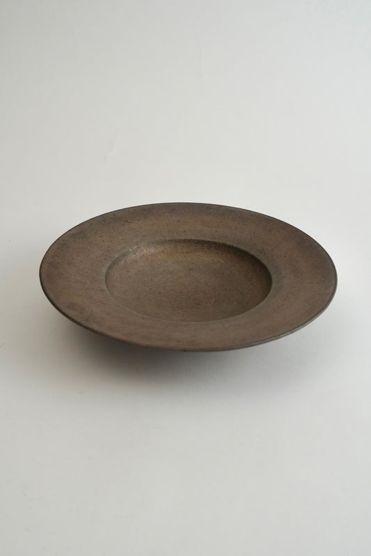 A round, shallow ceramic bowl with a wide rim, featuring a matte, dark brown textured glaze, set against a plain light gray background.