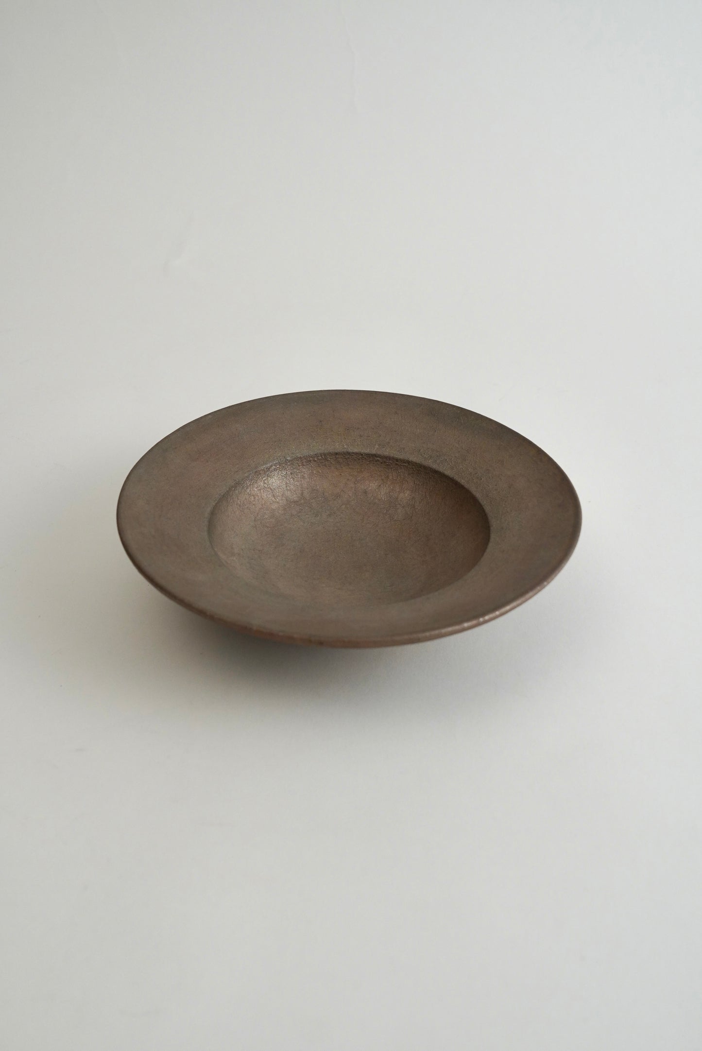 A shallow, round ceramic bowl with a matte, textured finish in a dark gray-brown color, photographed against a neutral light gray background.