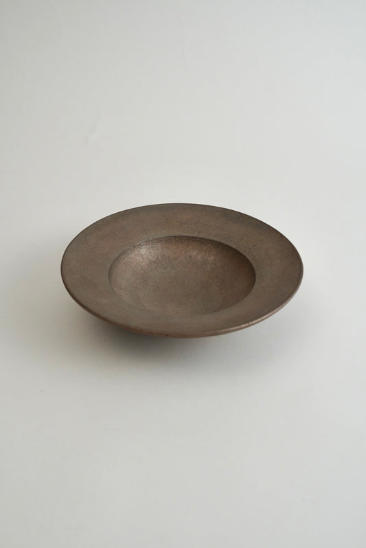 A shallow, round ceramic bowl with a matte, textured finish in a dark gray-brown color, photographed against a neutral light gray background.