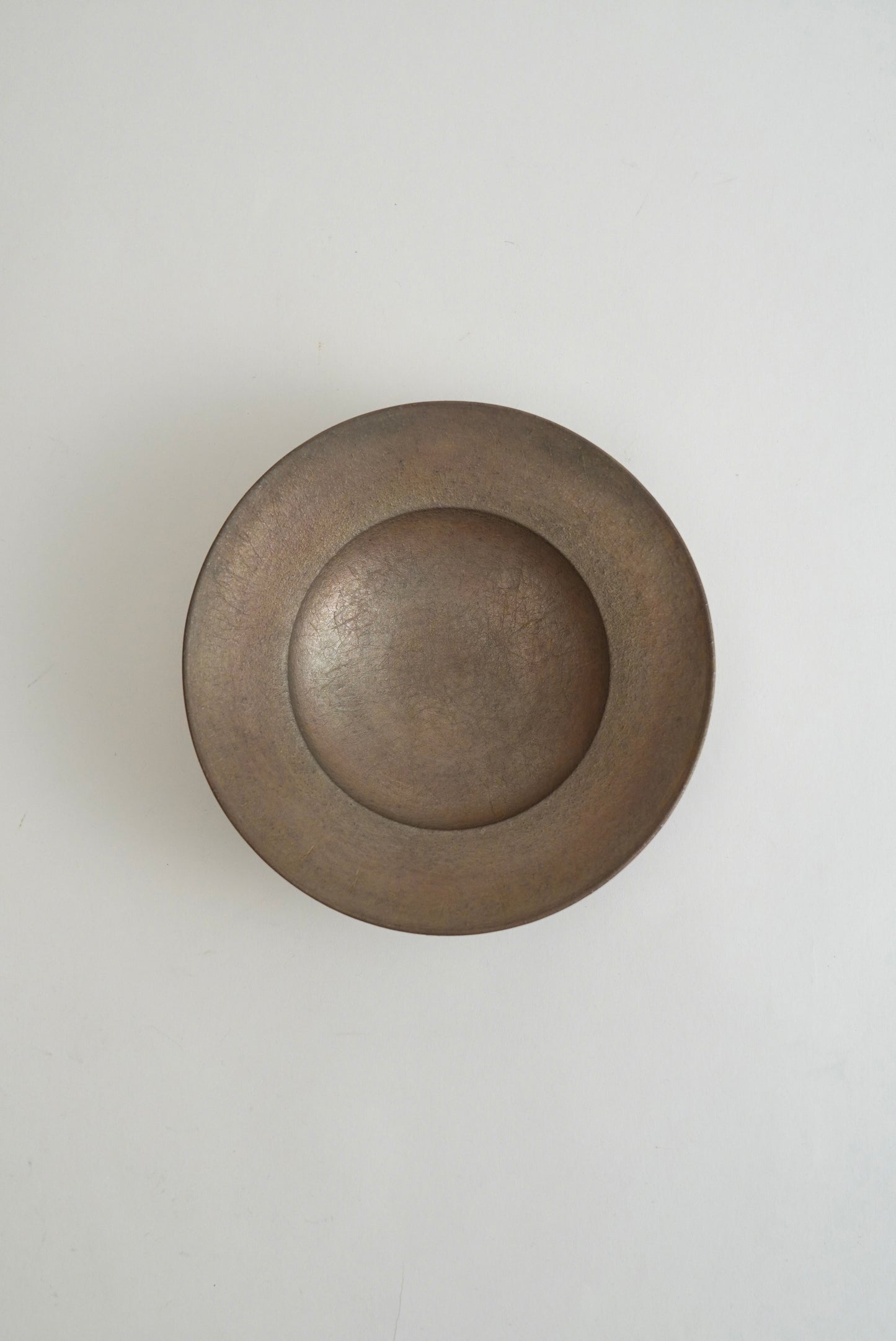 A round, shallow metal tray or dish with a textured, brushed finish in a warm bronze-brown color, shown from a top-down perspective on a plain light gray background.