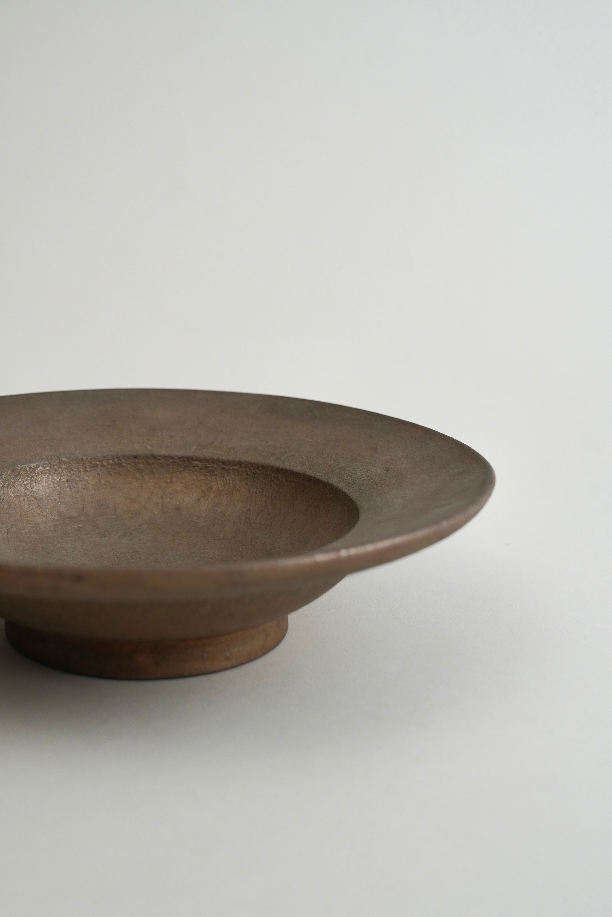A minimalist, shallow ceramic bowl with a wide rim and a small pedestal base, shown in a close-up side view against a plain light gray background.