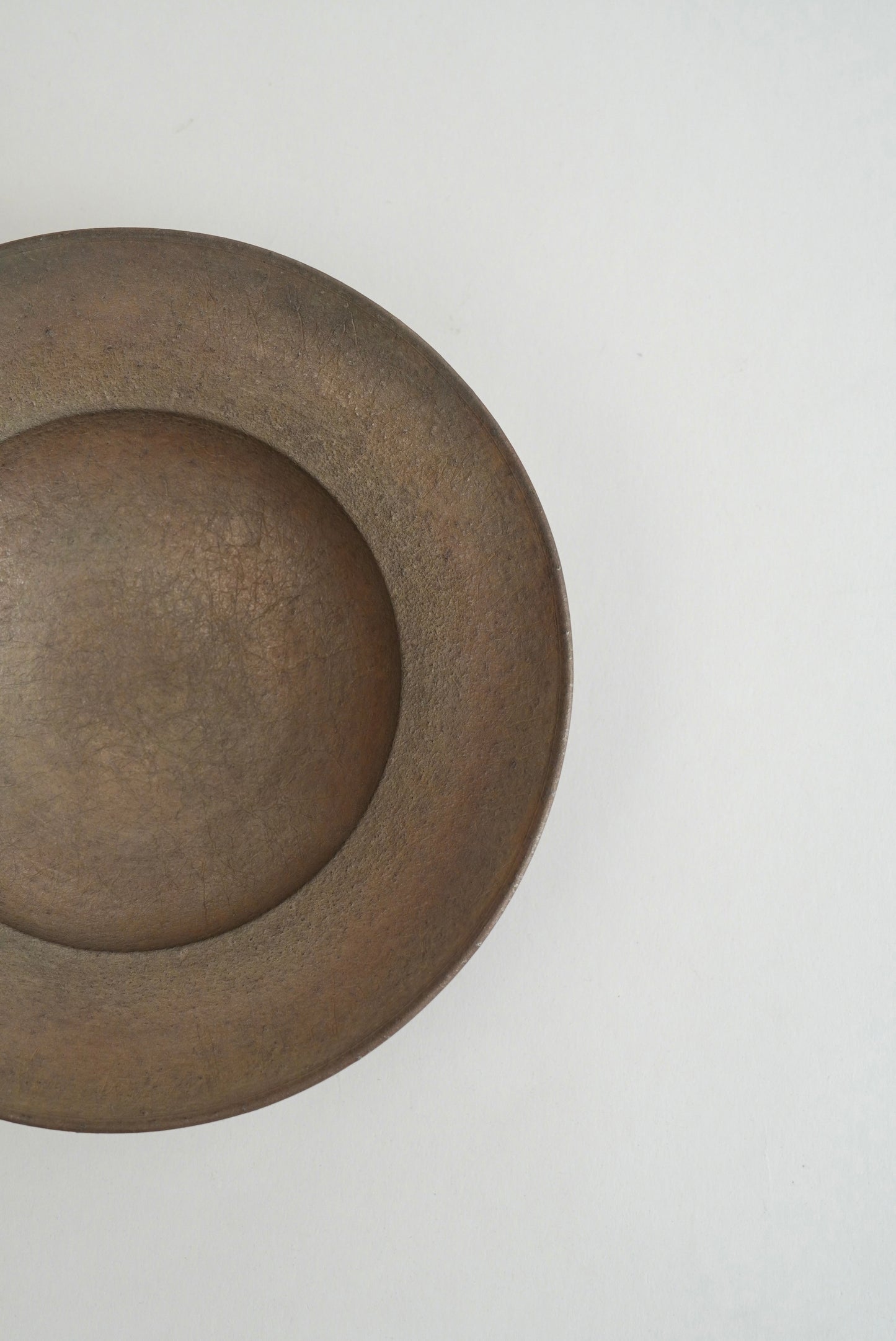 A close-up of a round, shallow bowl or dish with a matte, textured finish in a warm brownish-gray color, positioned on the left side of a plain light gray background.