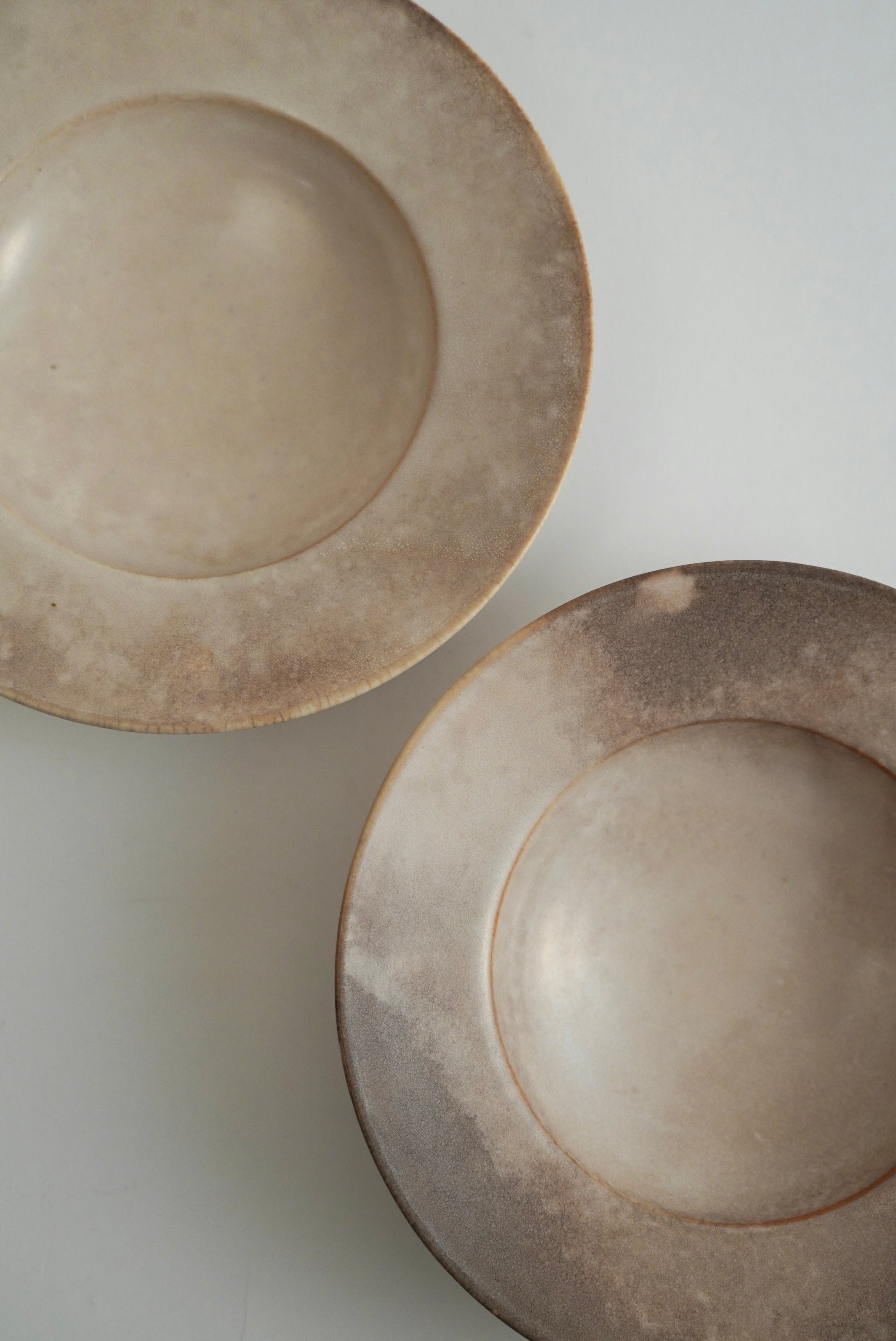 Two round ceramic plates with a rustic, textured matte finish in warm beige and gray tones, shown on a light gray background.
