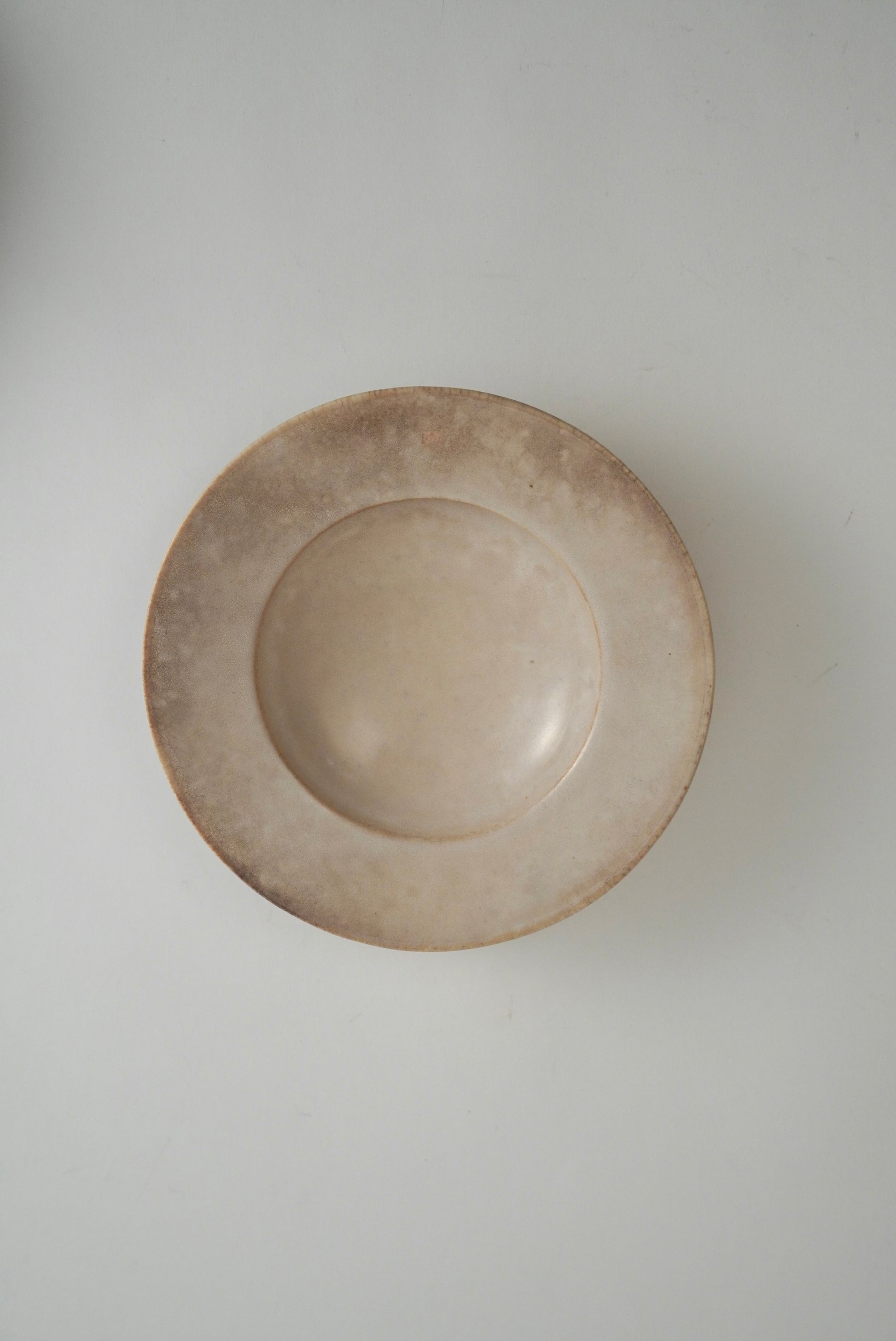 A round, shallow ceramic plate or dish with a rustic, hand-painted finish in a light beige and brown glaze, shown from a top-down perspective on a neutral light gray background.