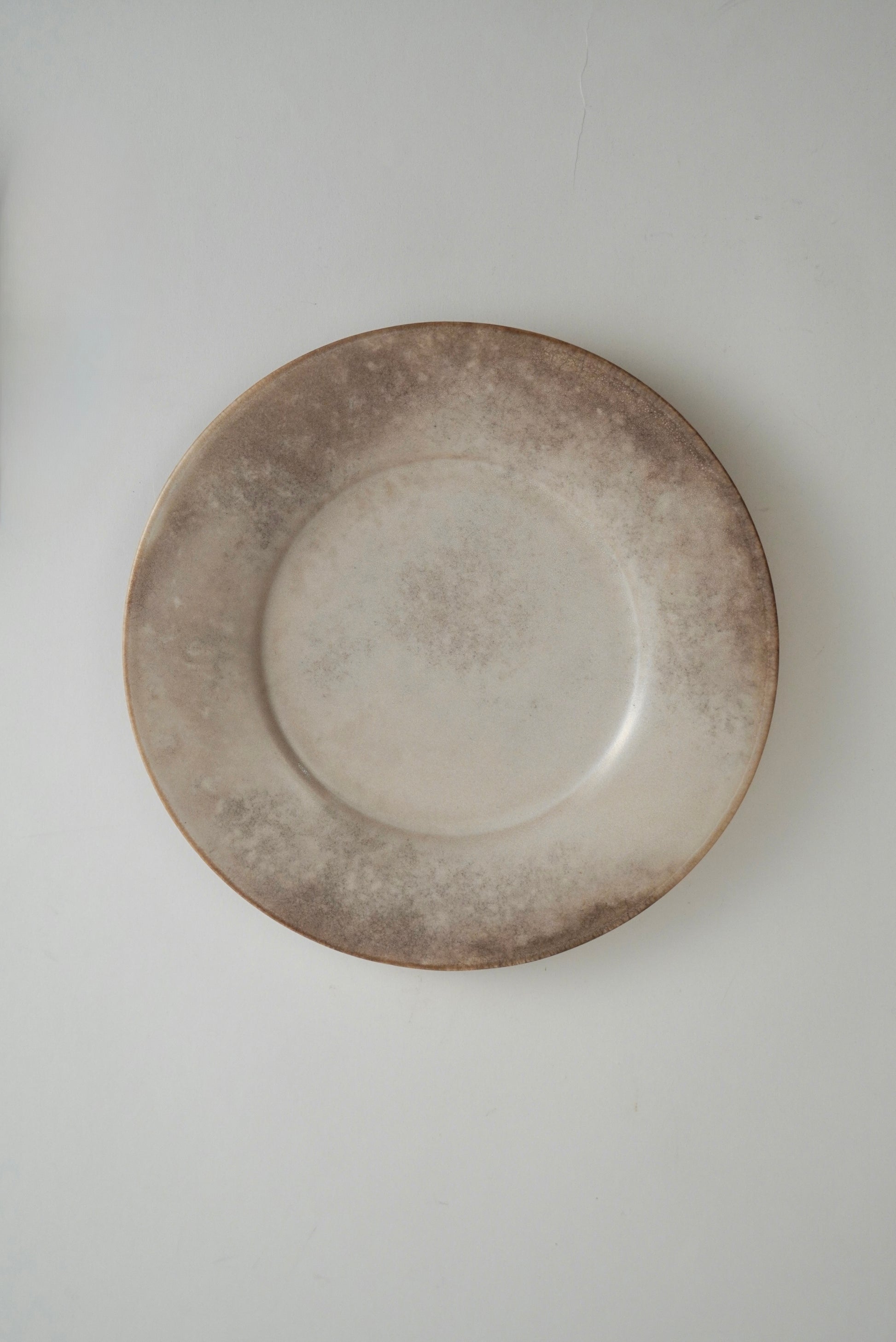 A round ceramic plate with a rustic, distressed finish, featuring a light beige center and a mottled, darker brown rim, shown against a plain light gray background.