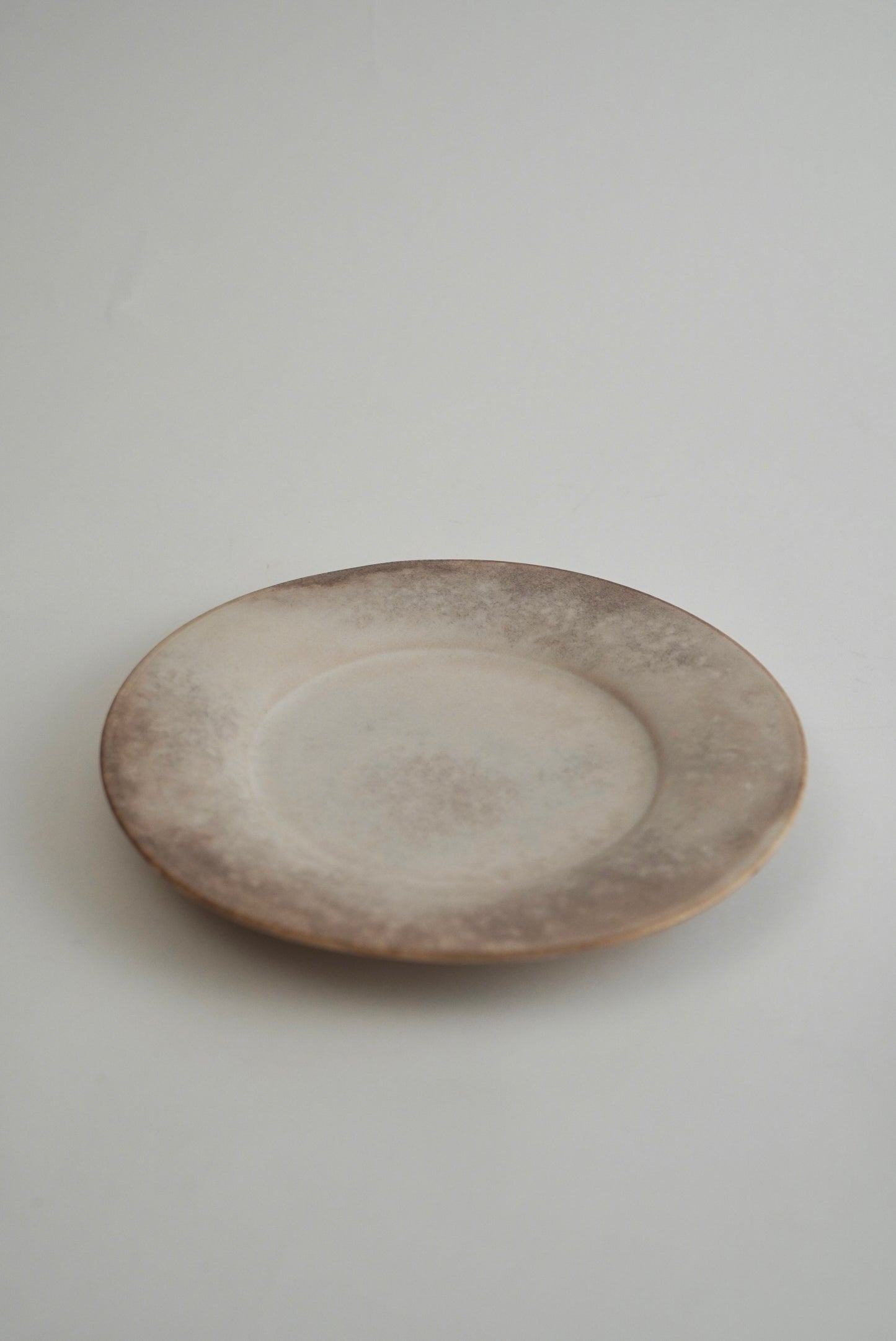 A round ceramic plate with a rustic, distressed finish in earthy brown and beige tones, photographed on a neutral light gray background.