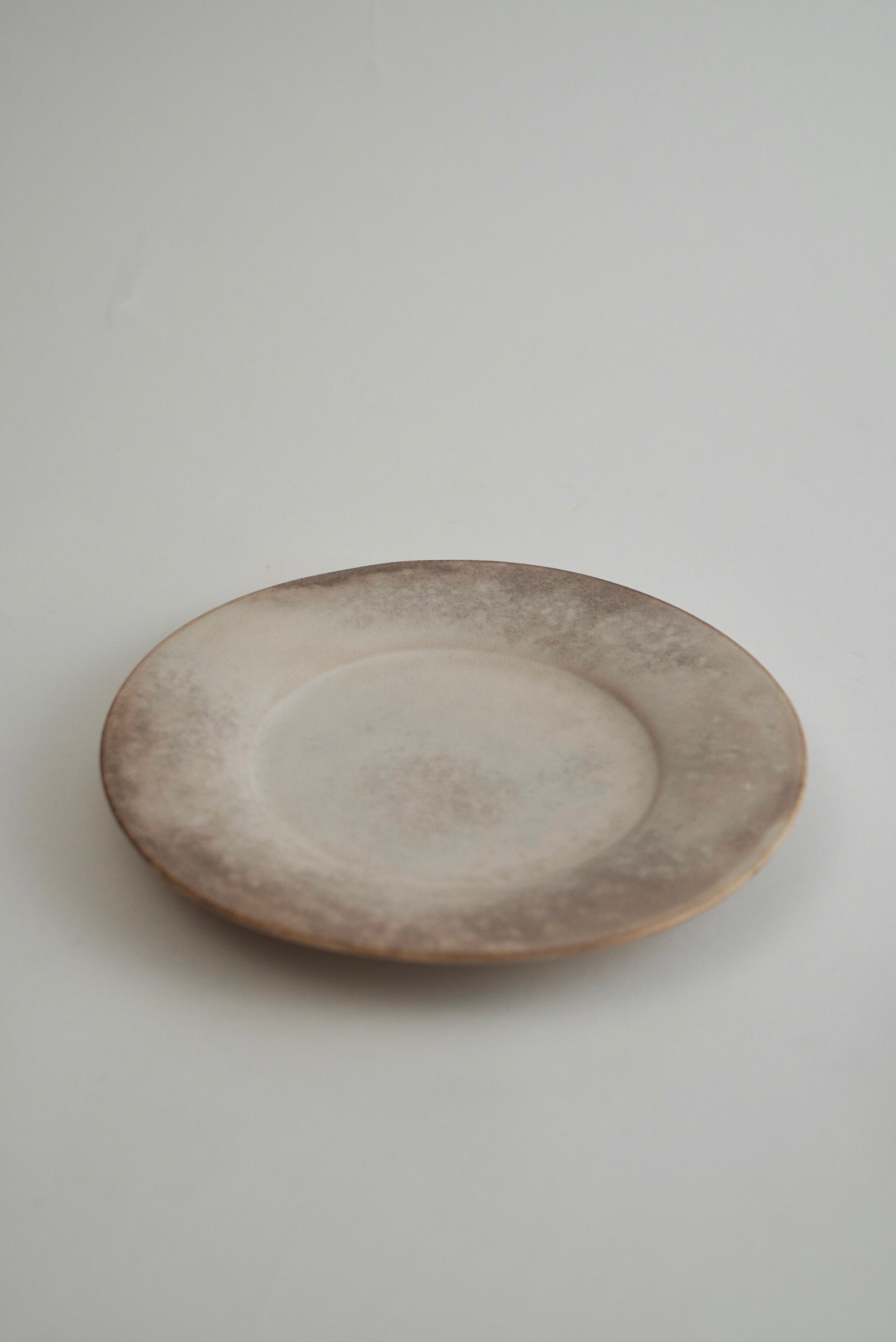 A round ceramic plate with a rustic, distressed finish in earthy brown and beige tones, photographed on a neutral light gray background.