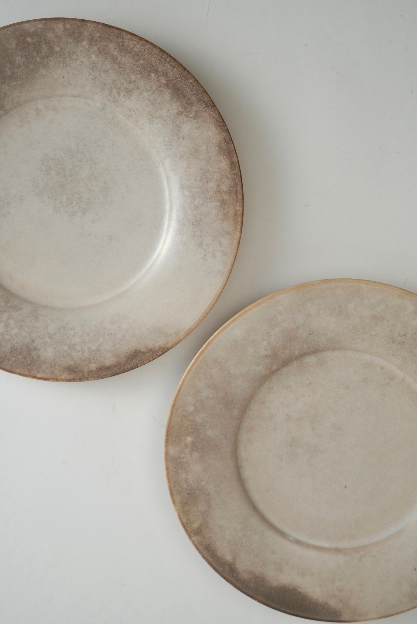 Two round ceramic plates with a distressed, mottled beige and brown glaze, shown on a plain white background.