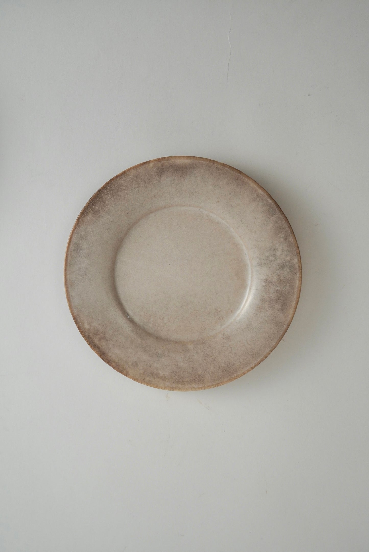 A round ceramic plate with a rustic, hand-painted finish in earthy beige and brown tones, photographed from directly above against a neutral light gray background.