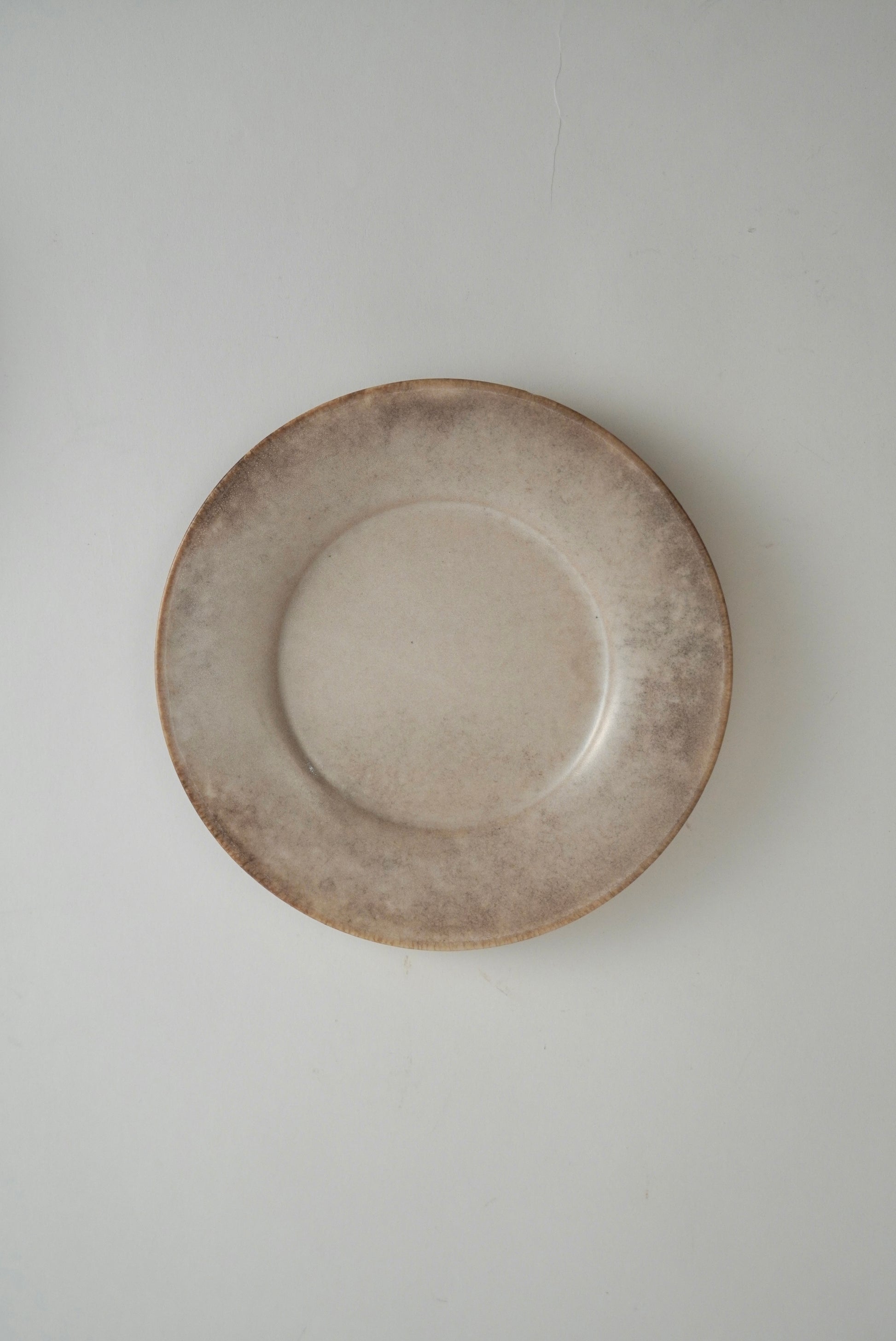 A round ceramic plate with a rustic, hand-painted finish in earthy beige and brown tones, photographed from directly above against a neutral light gray background.