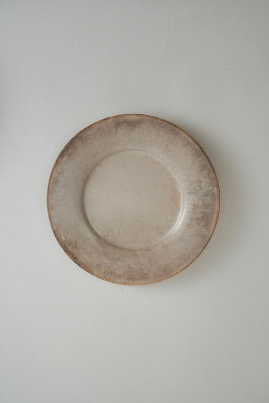 A round ceramic plate with a rustic, hand-painted finish in earthy beige and brown tones, photographed from directly above against a neutral light gray background.