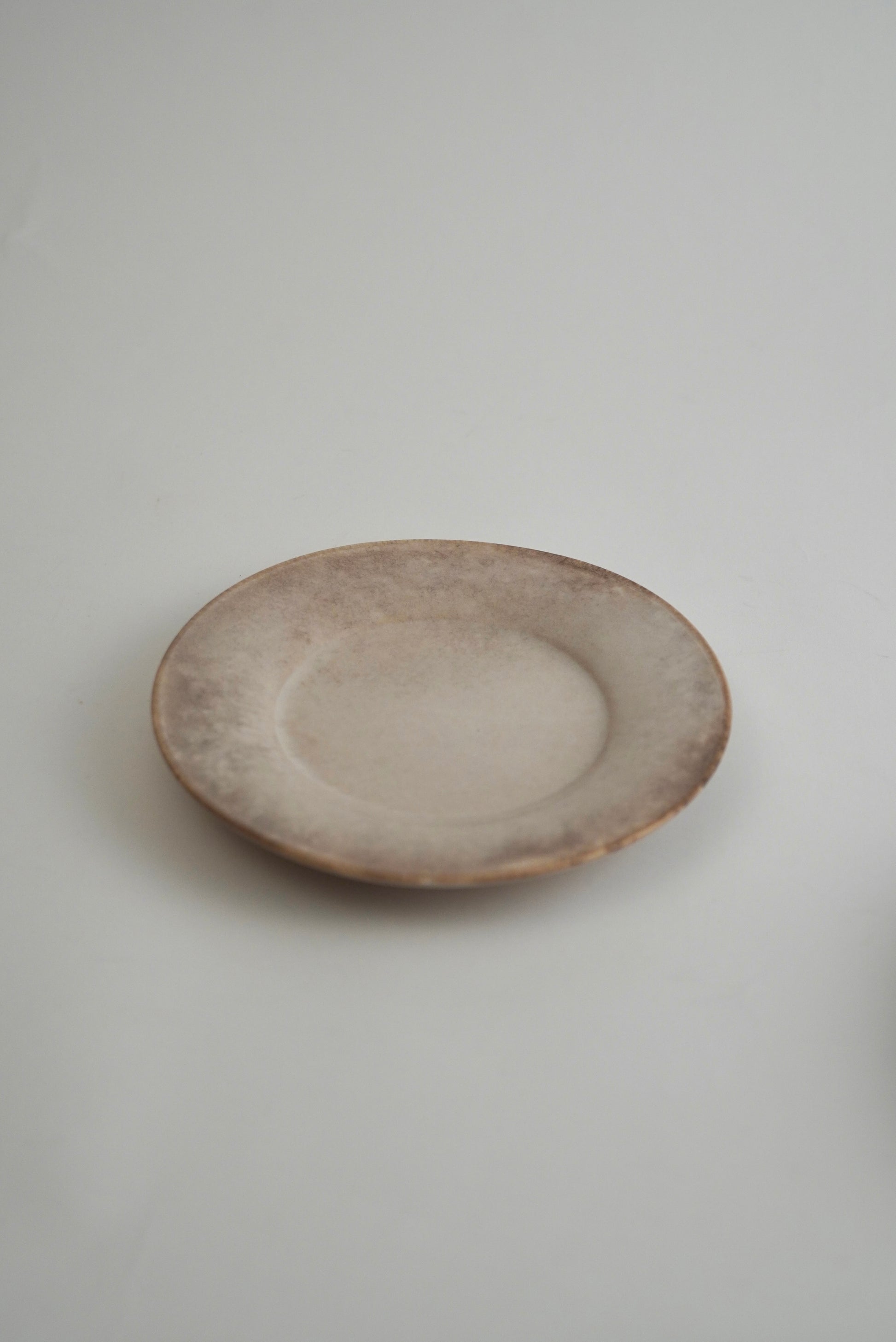 A small round ceramic plate with a rustic, hand-painted finish in muted beige and brown tones, set against a plain light gray background.