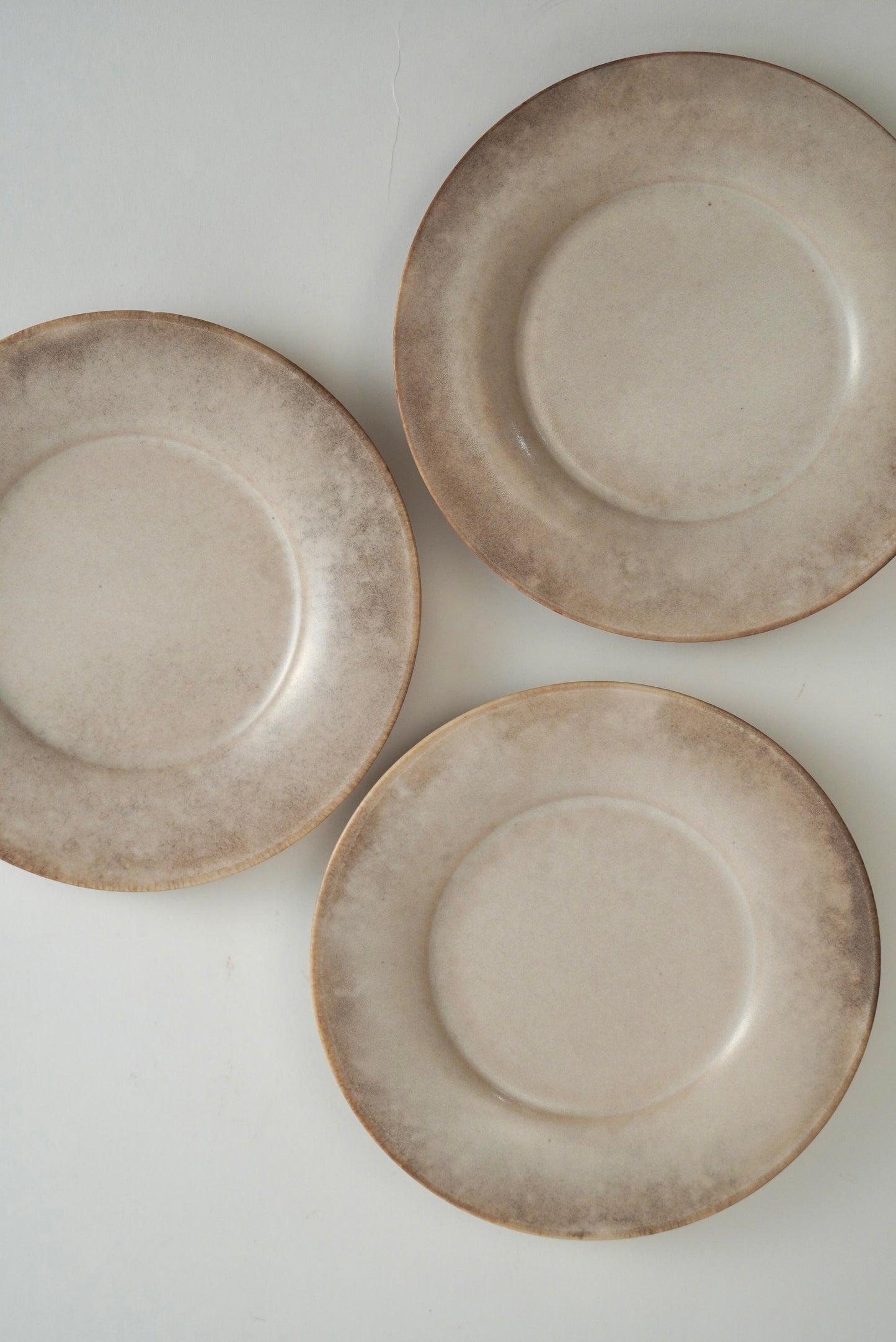 Three round ceramic plates with a distressed, mottled beige and brown glaze, arranged on a plain white background.