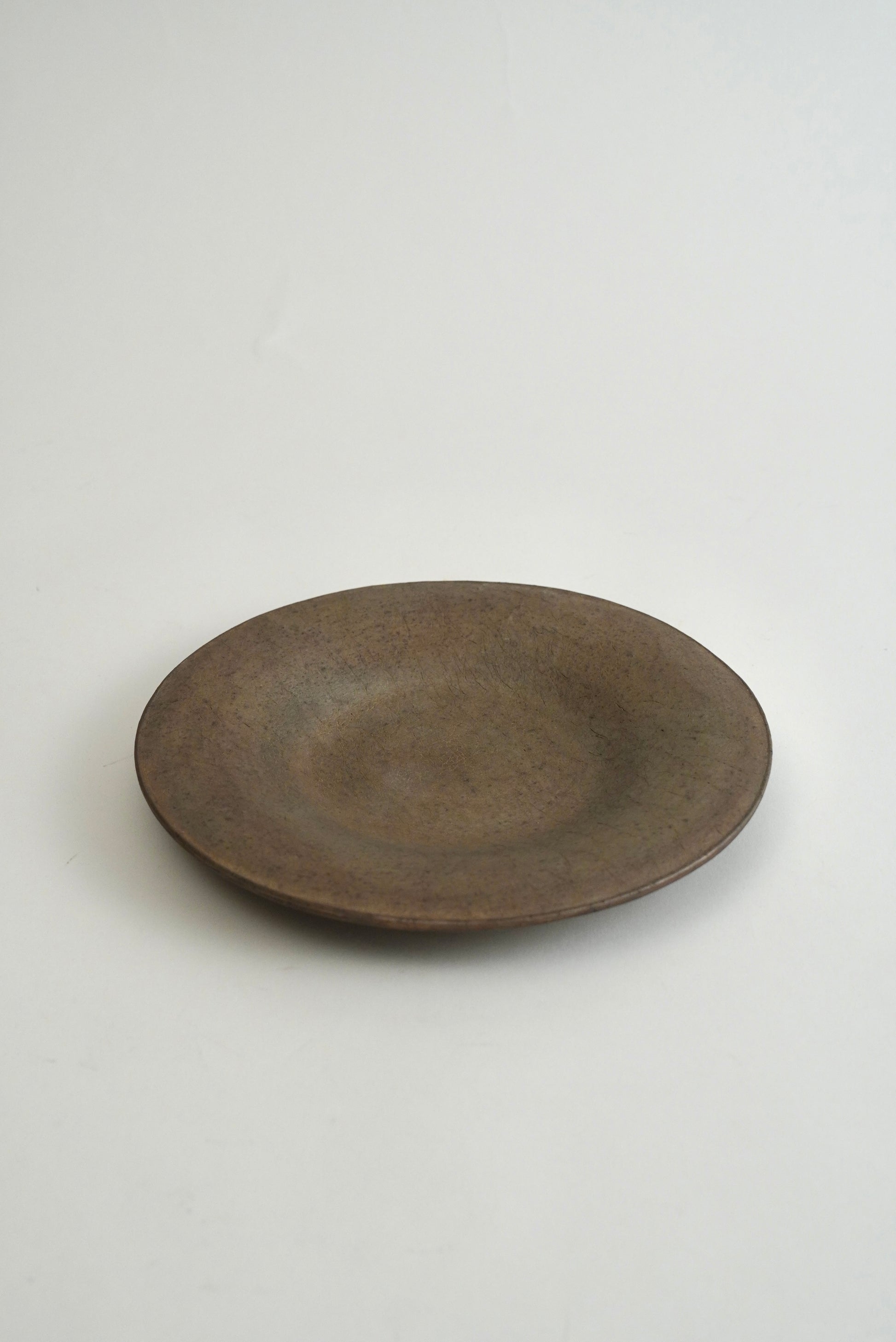 A round, shallow ceramic plate with a rustic, matte dark brown finish, photographed against a plain light gray background.