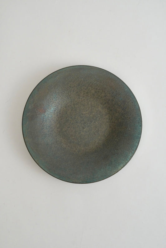 A round ceramic plate with a textured, speckled glaze in dark gray and muted green tones, photographed from directly above on a light gray background.