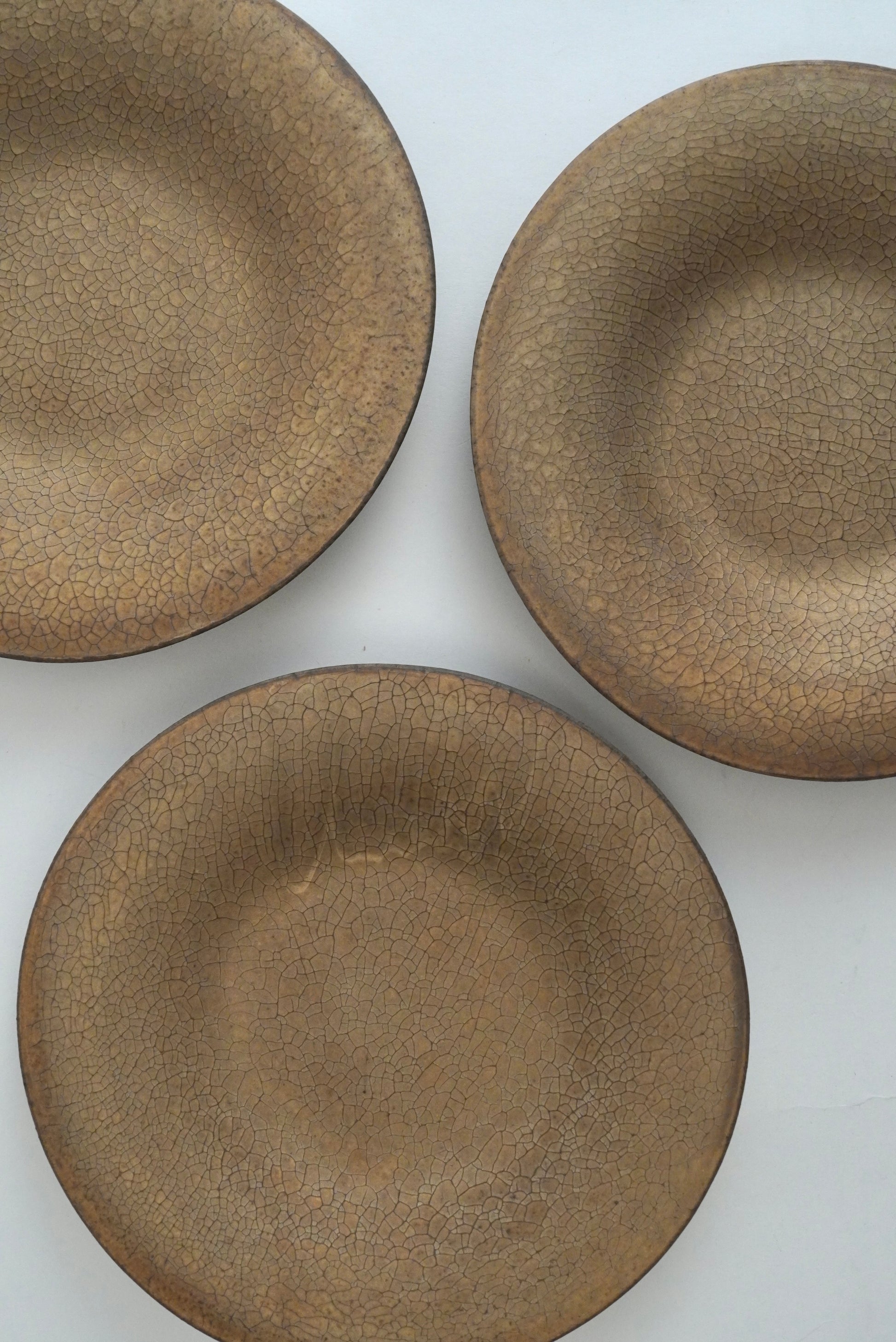 Three round ceramic plates with a textured, crackled glaze in a warm taupe-brown color, arranged on a plain white background.