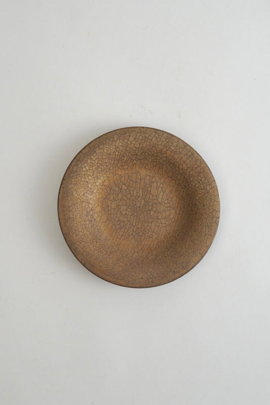 A round ceramic plate with a textured, crackled glaze in a warm brown and beige color, photographed from directly above on a light gray background.