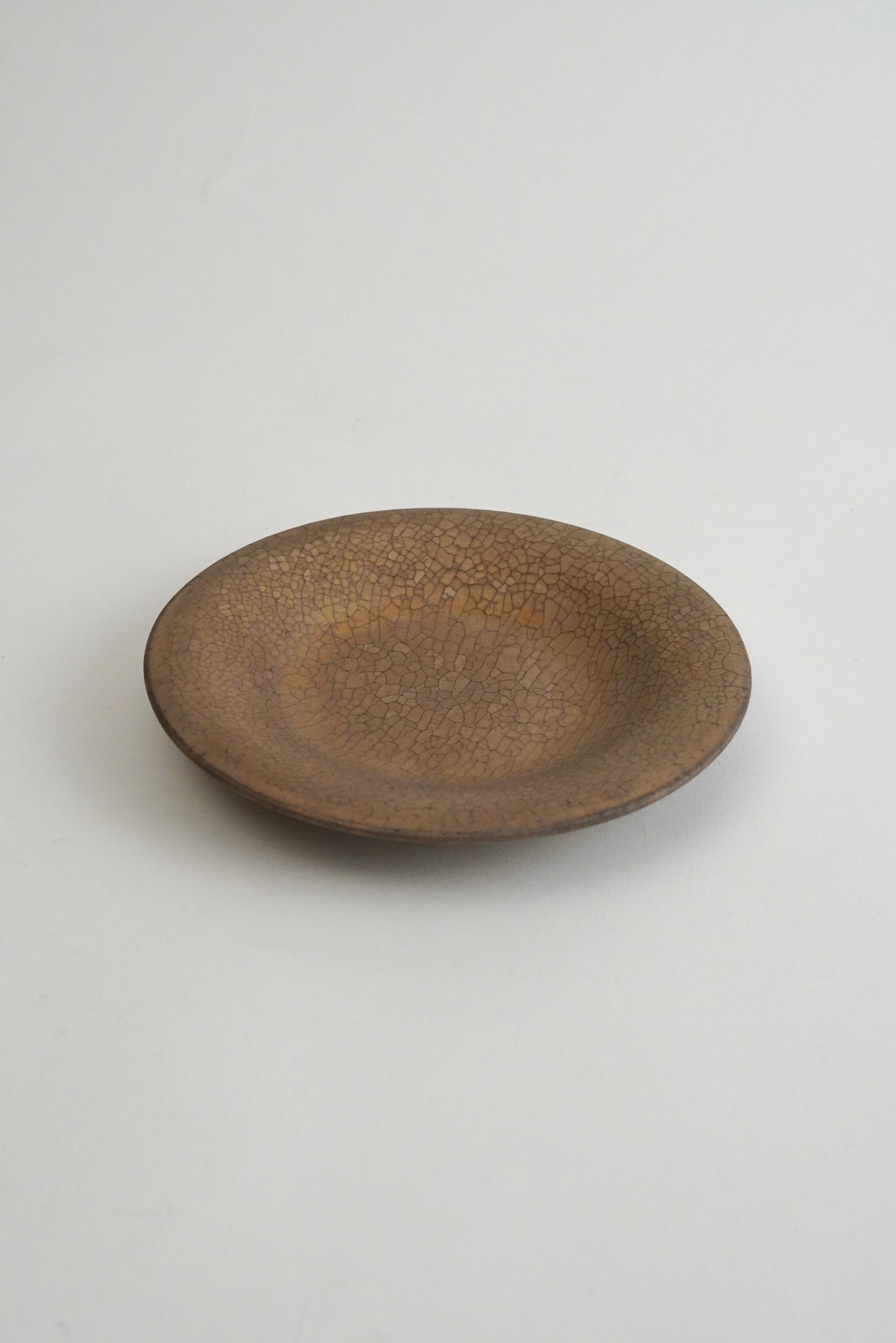 A small, round ceramic plate with a textured, speckled brown glaze, photographed from a top-down angle on a neutral light gray background.