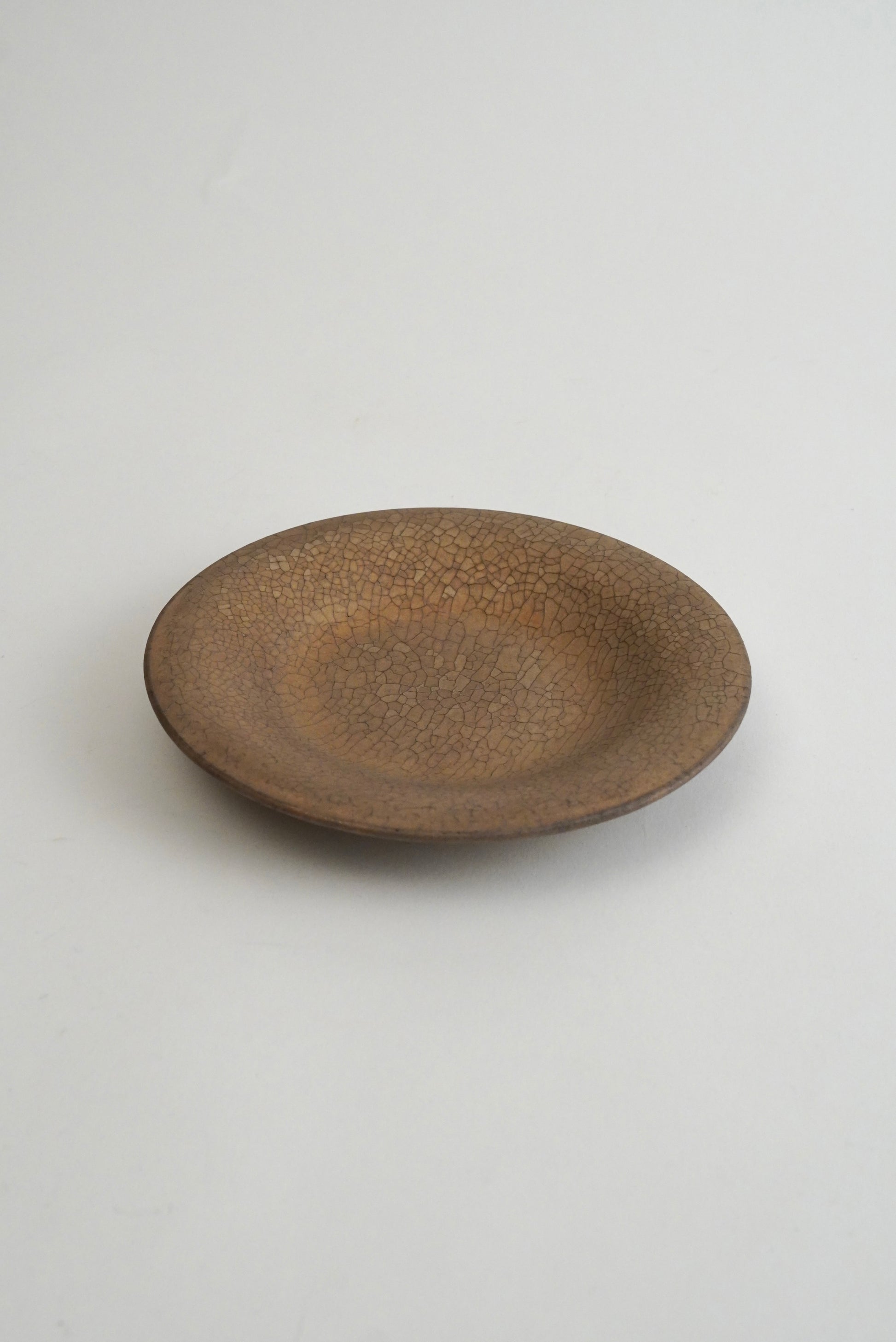 A small, round ceramic plate with a textured, speckled brown glaze, photographed from a top-down angle on a neutral light gray background.