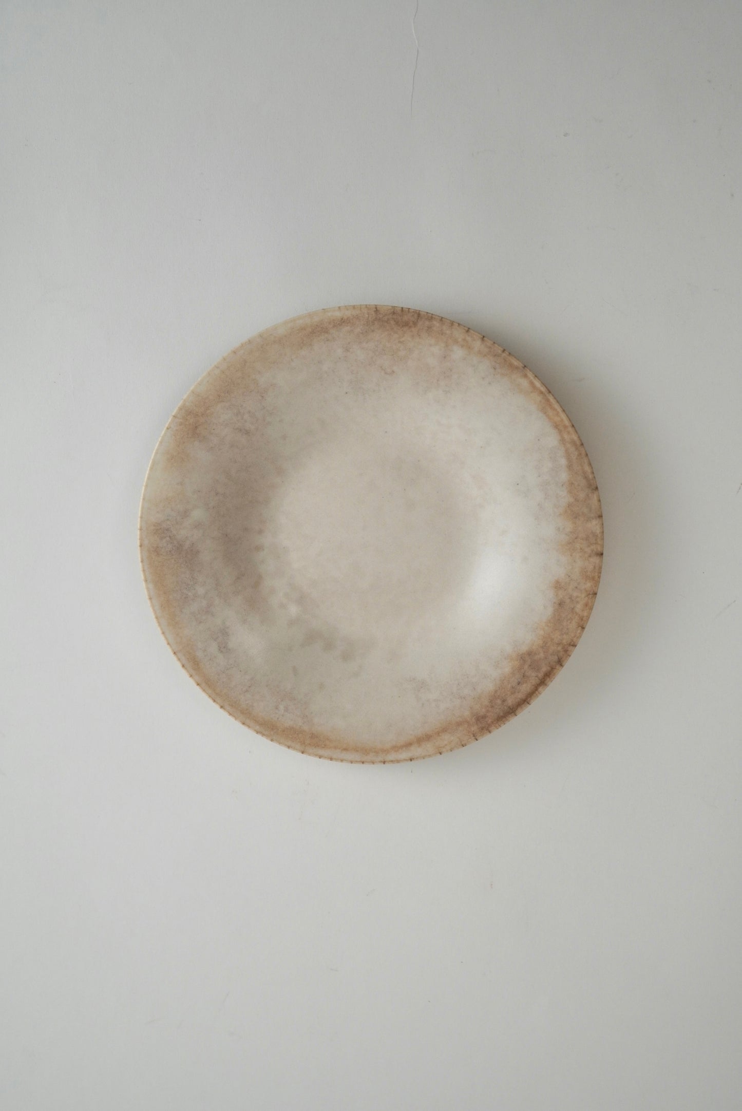 A round ceramic plate with a textured, rustic finish in shades of beige and light brown, photographed from directly above on a neutral light gray background.