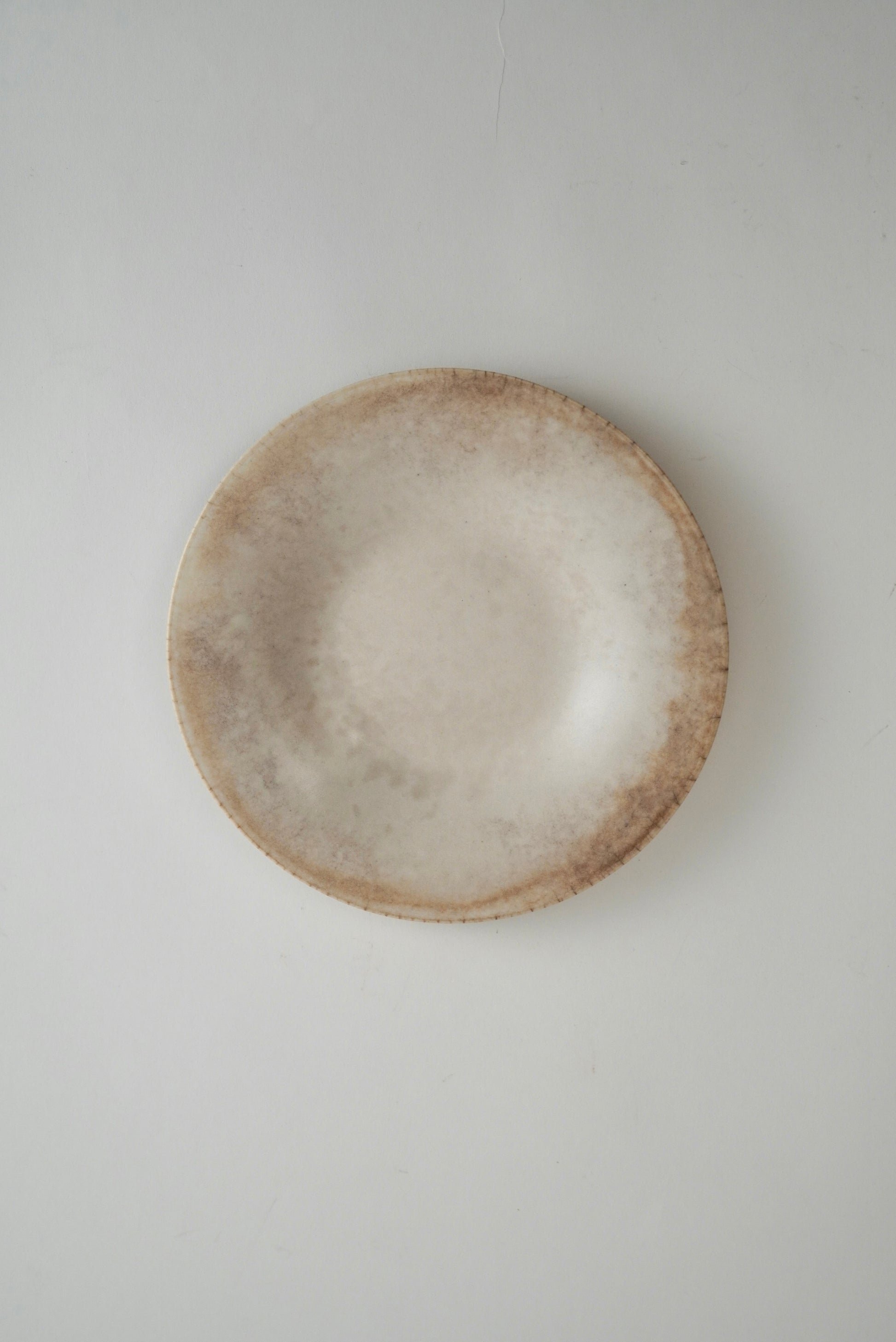 A round ceramic plate with a textured, rustic finish in shades of beige and light brown, photographed from directly above on a neutral light gray background.
