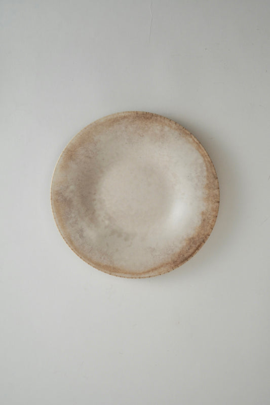 A round ceramic plate with a textured, rustic finish in shades of beige and light brown, photographed from directly above on a neutral light gray background.