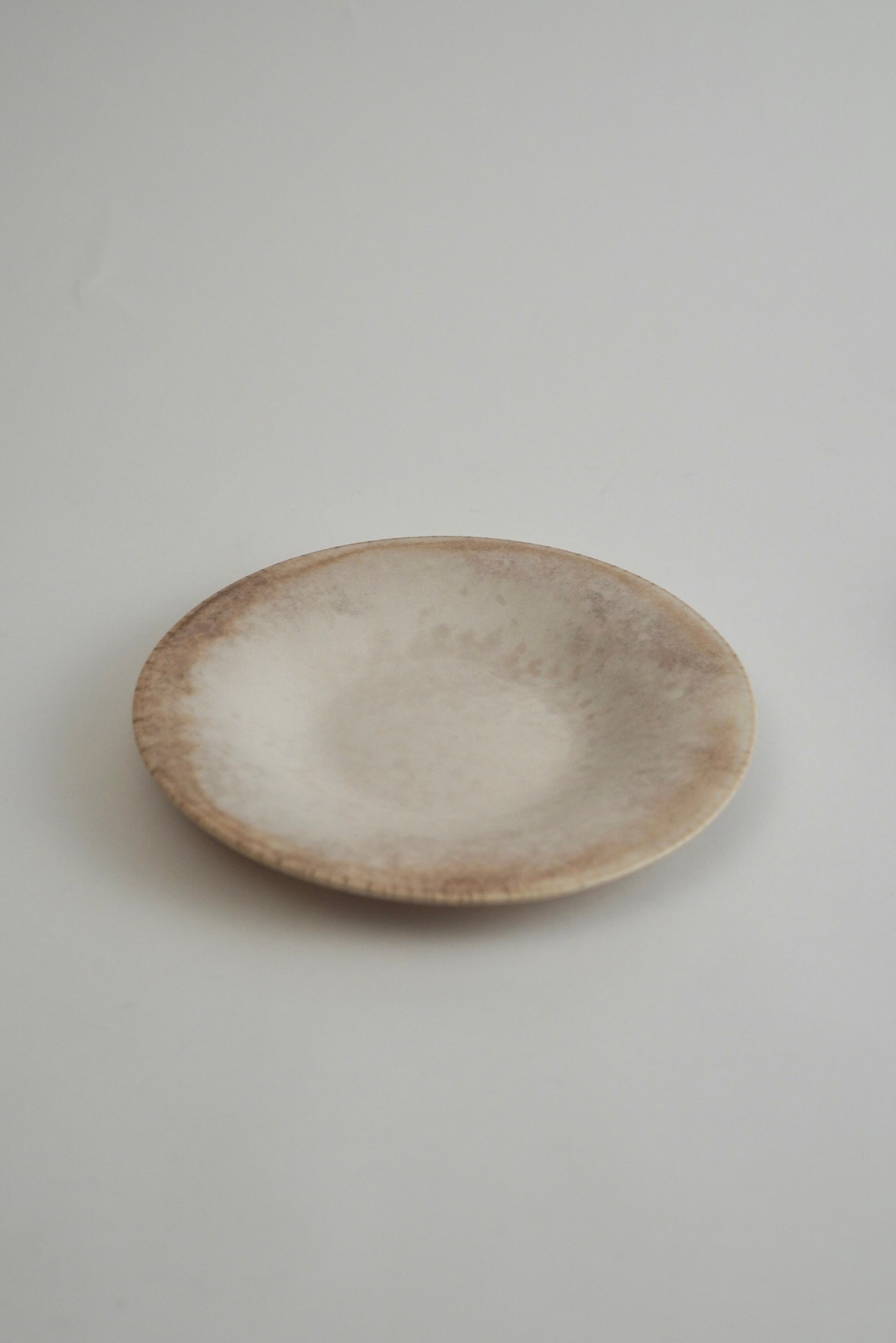 A small round ceramic plate with a textured, mottled finish in shades of beige, cream, and light brown, photographed on a neutral light gray background.