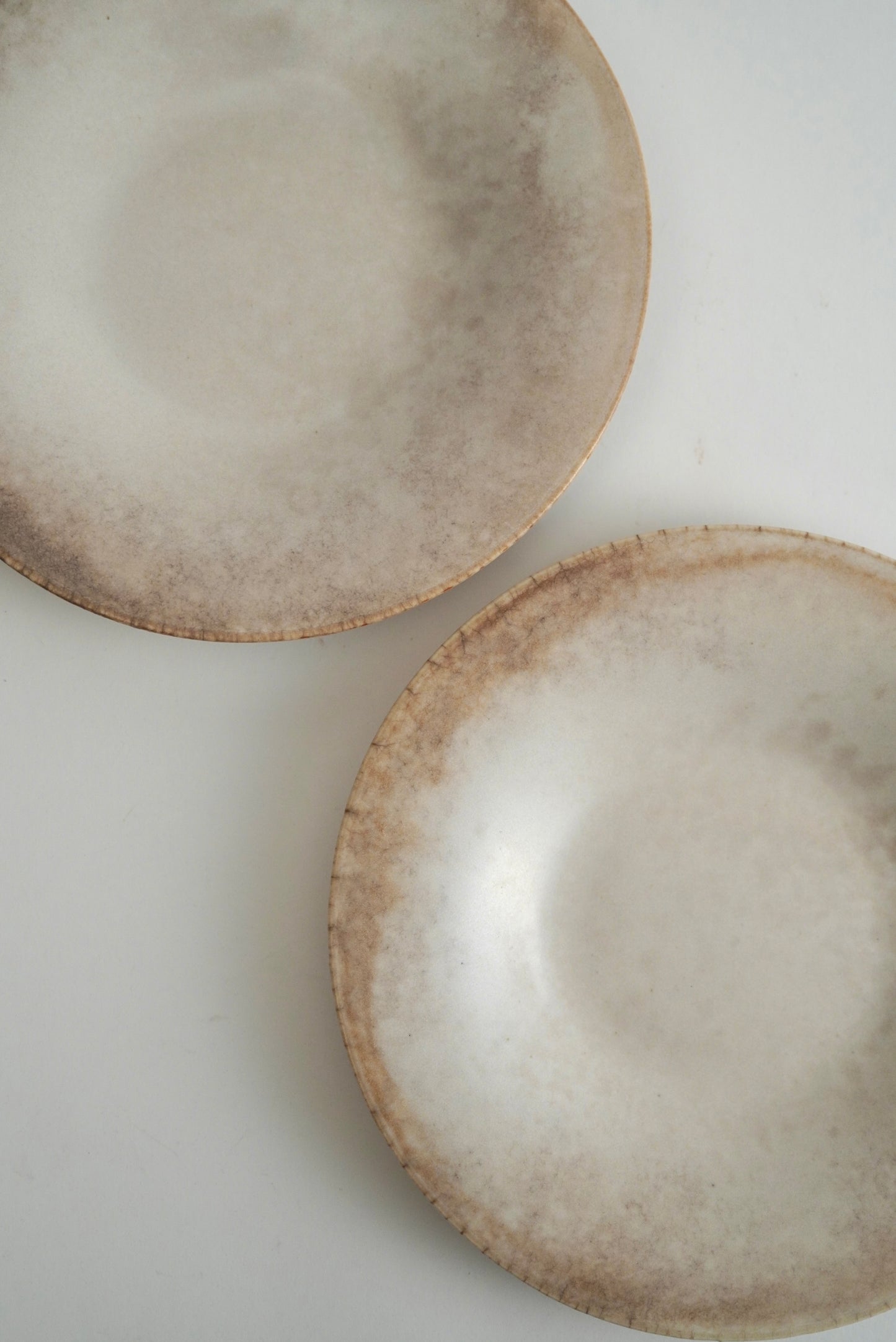 Two round ceramic plates with a textured, rustic finish in warm beige and brown tones, photographed on a plain white surface.