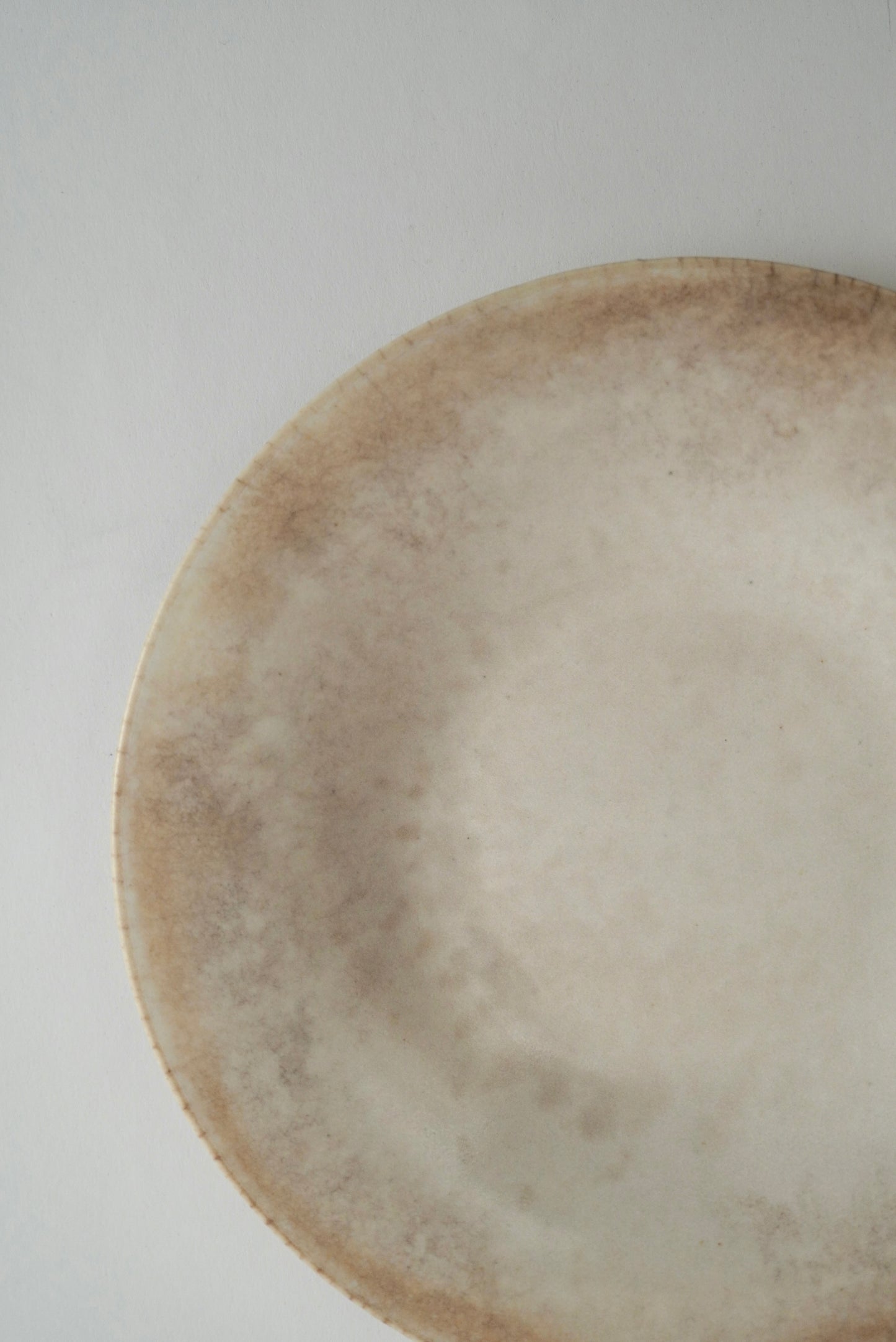 A close-up of a round ceramic plate with a rustic, hand-painted finish in earthy beige and brown tones, set against a plain light gray background.