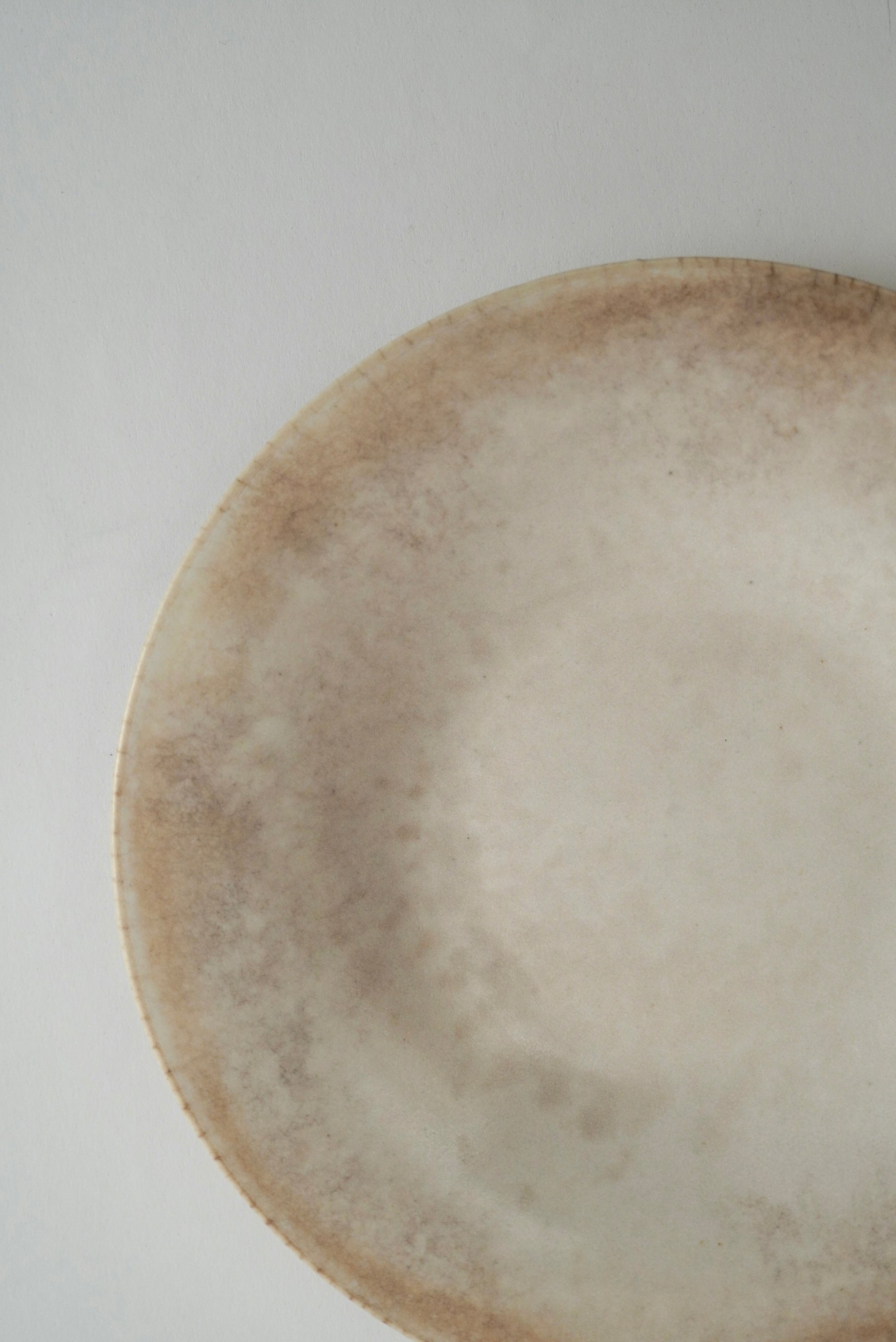 A close-up of a round ceramic plate with a rustic, hand-painted finish in earthy beige and brown tones, set against a plain light gray background.