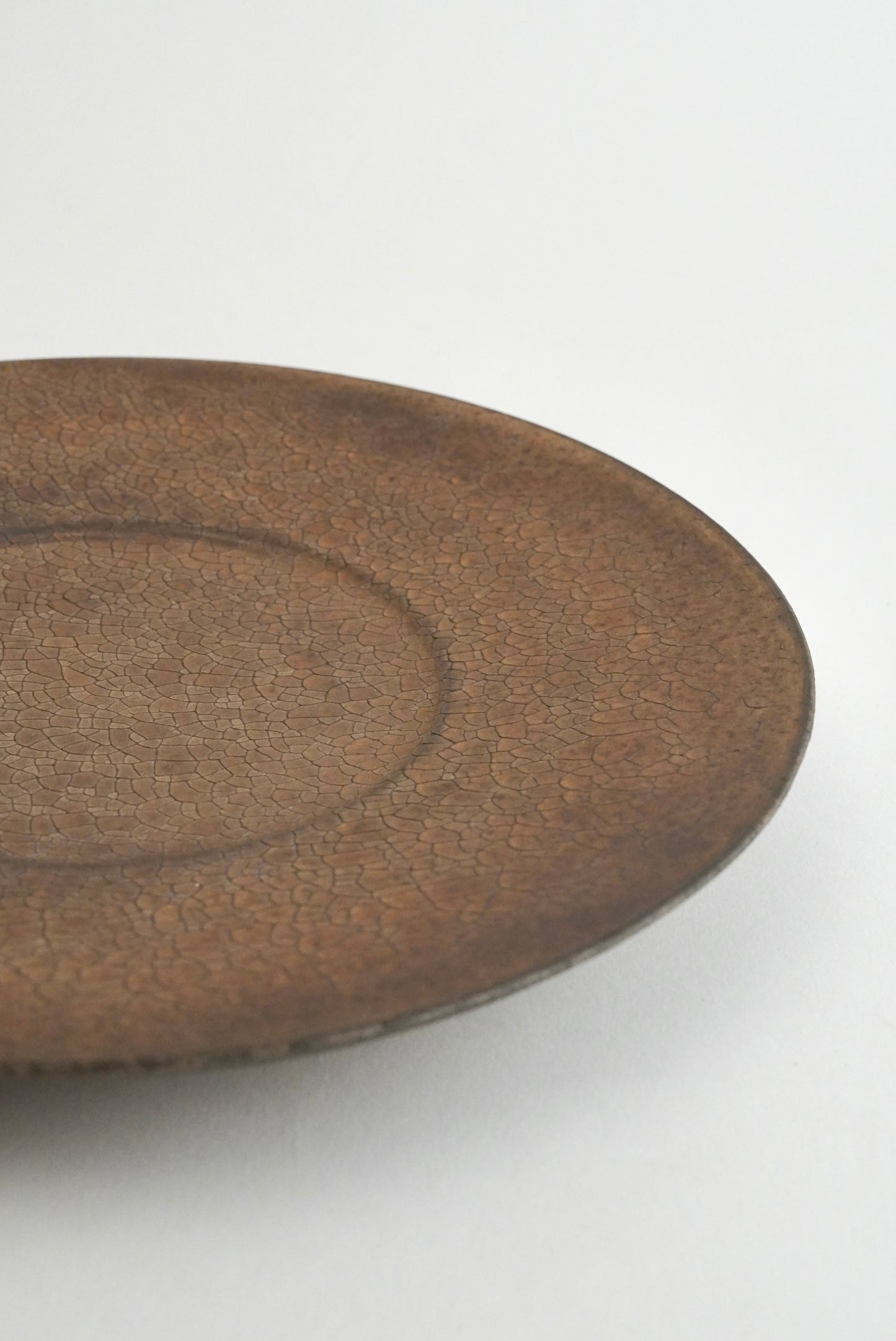 A close-up of a round ceramic plate with a textured, earthy brown glaze on a plain white background.