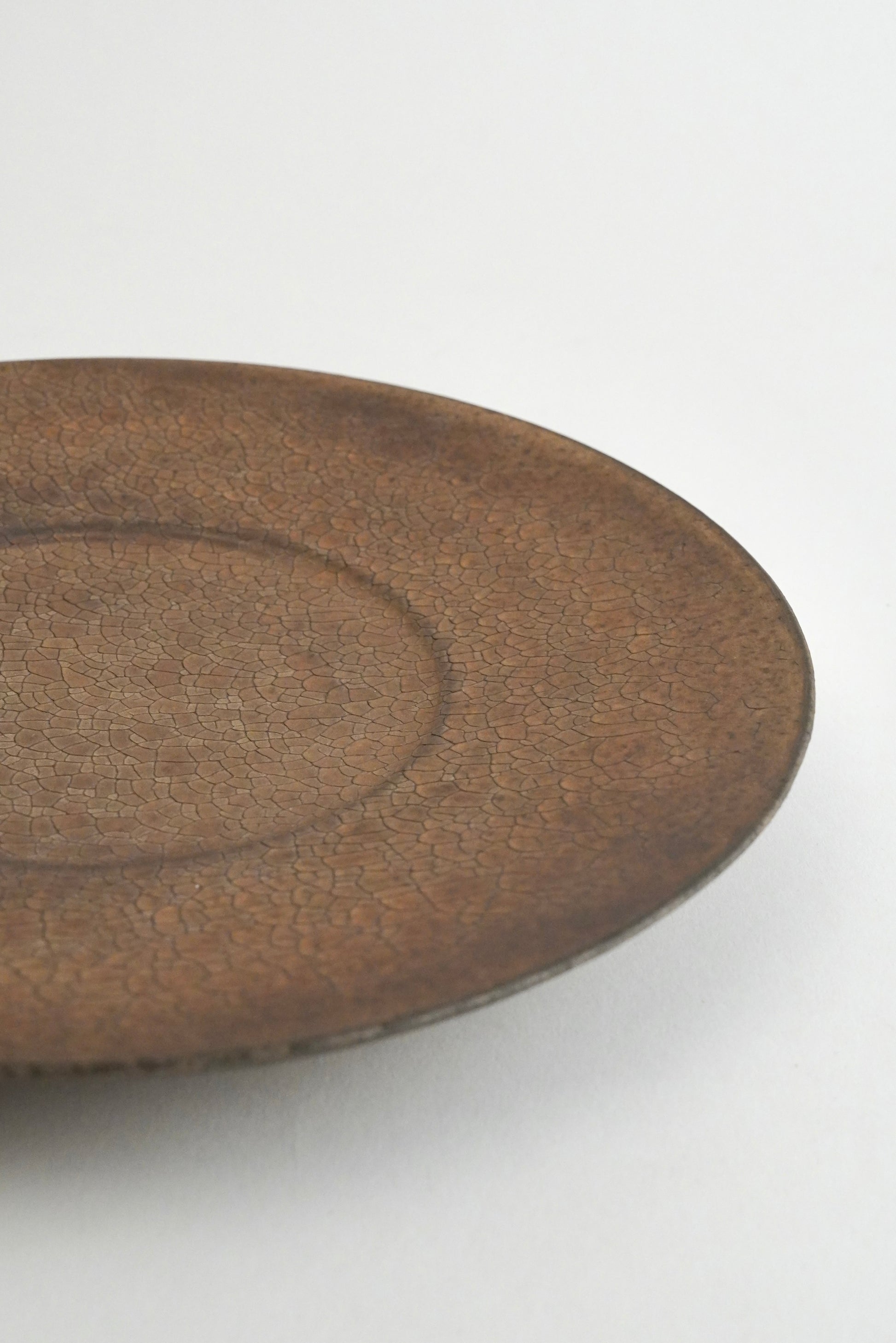 A close-up of a round ceramic plate with a textured, earthy brown glaze on a plain white background.