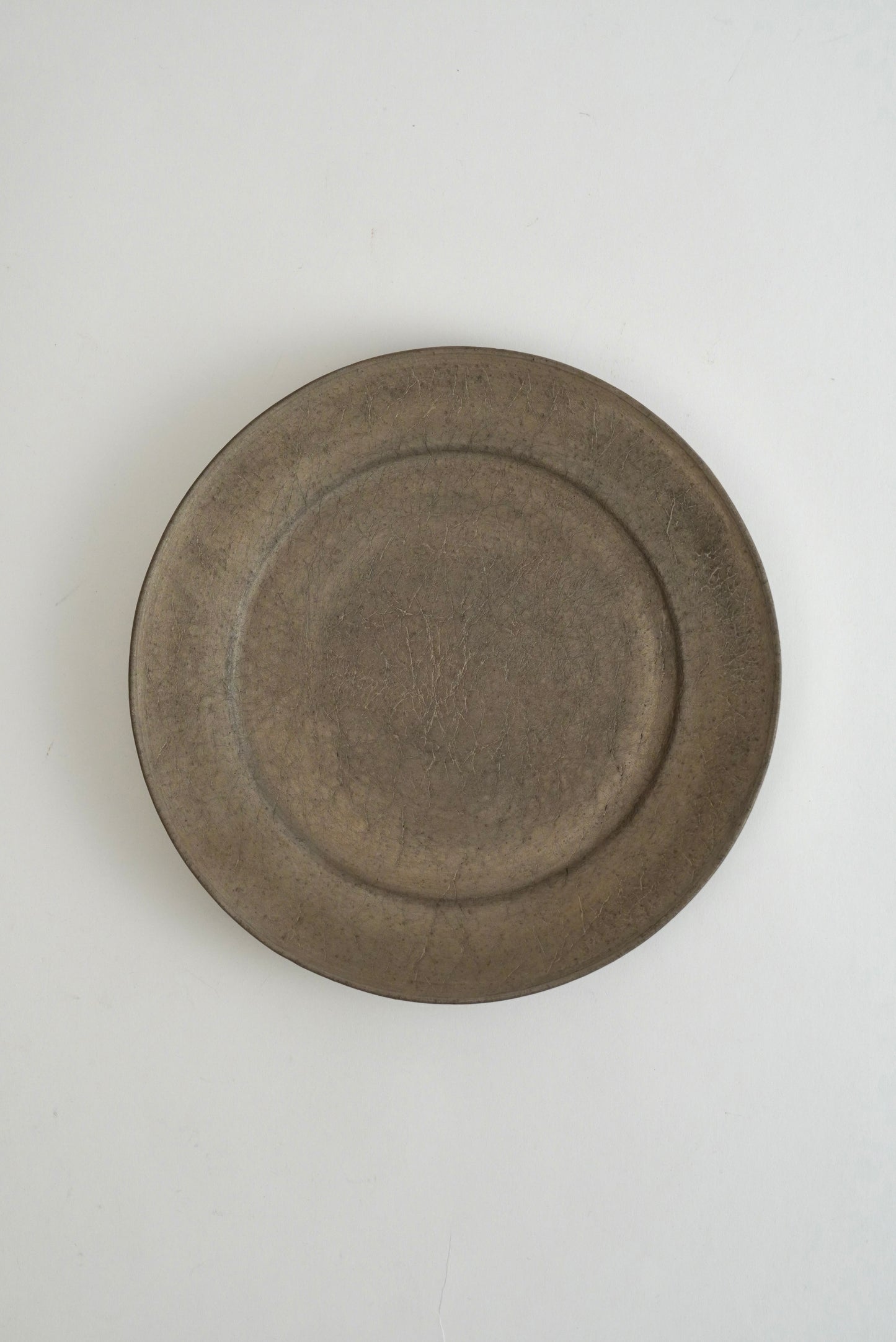 A round, shallow ceramic plate with a rustic, matte finish in a muted brownish-gray color, photographed from directly above on a light off-white background.