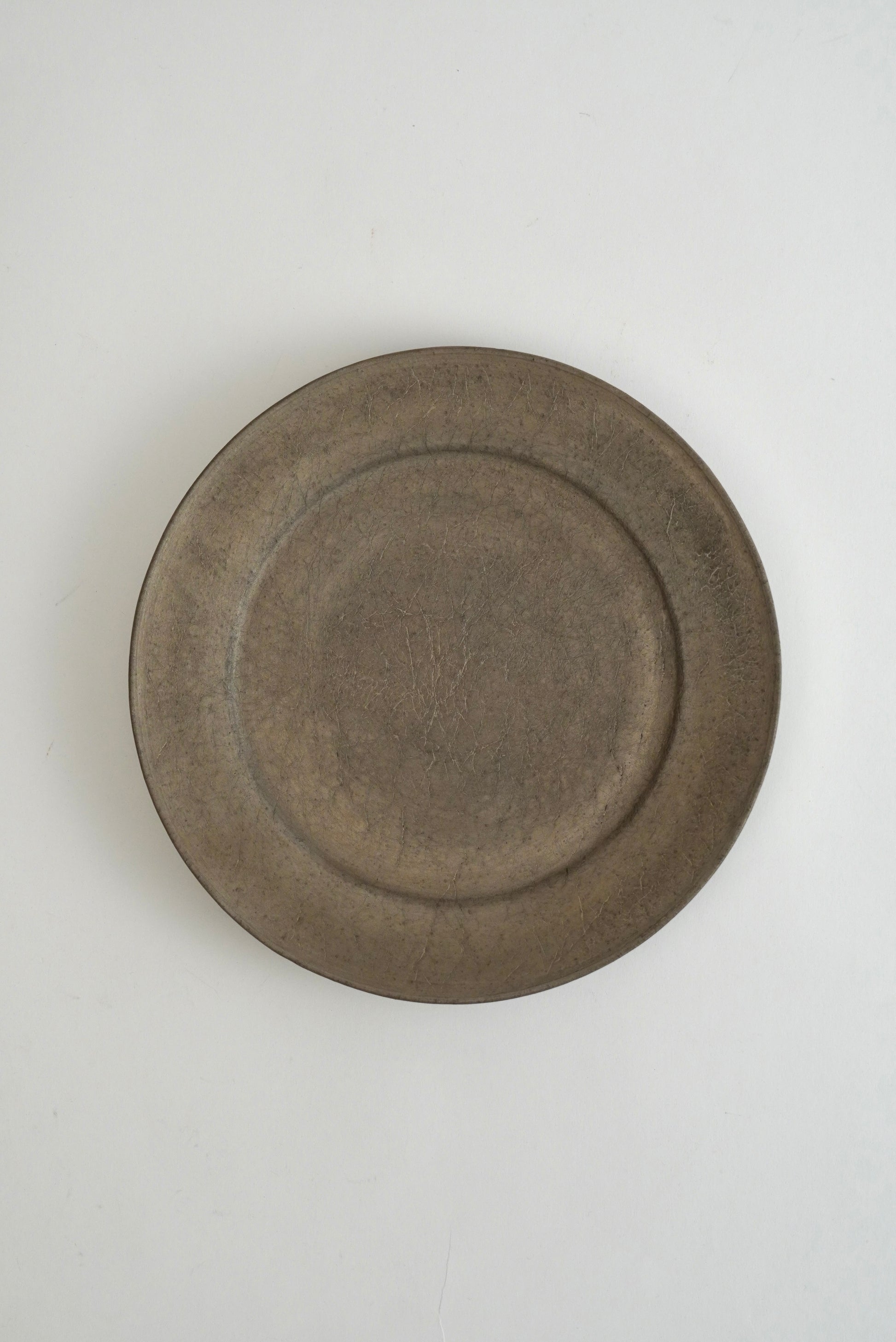 A round, shallow ceramic plate with a rustic, matte finish in a muted brownish-gray color, photographed from directly above on a light off-white background.