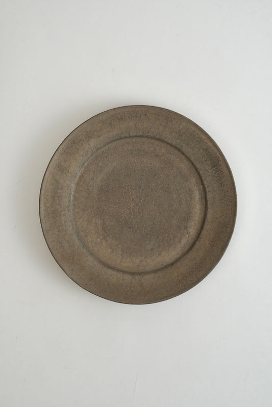 A round, shallow ceramic plate with a rustic, matte finish in a muted brownish-gray color, photographed from directly above on a light off-white background.