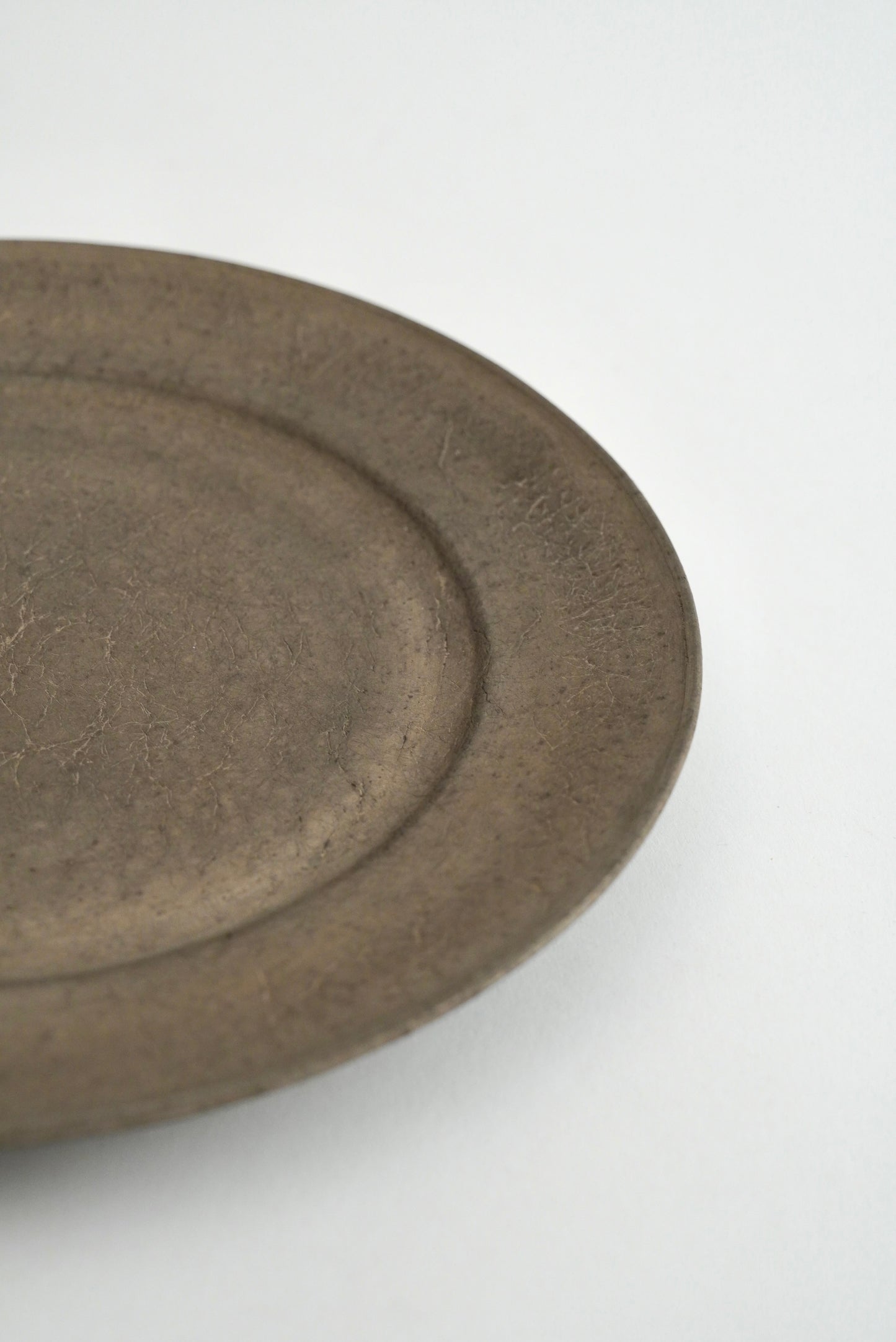A close-up of a round, textured ceramic plate or dish in a matte dark gray-brown glaze, shown on a plain white background.