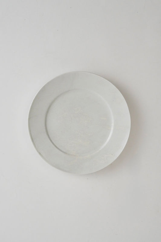 A minimalist white ceramic plate with a subtle textured pattern, photographed from directly above on a clean, light gray background.