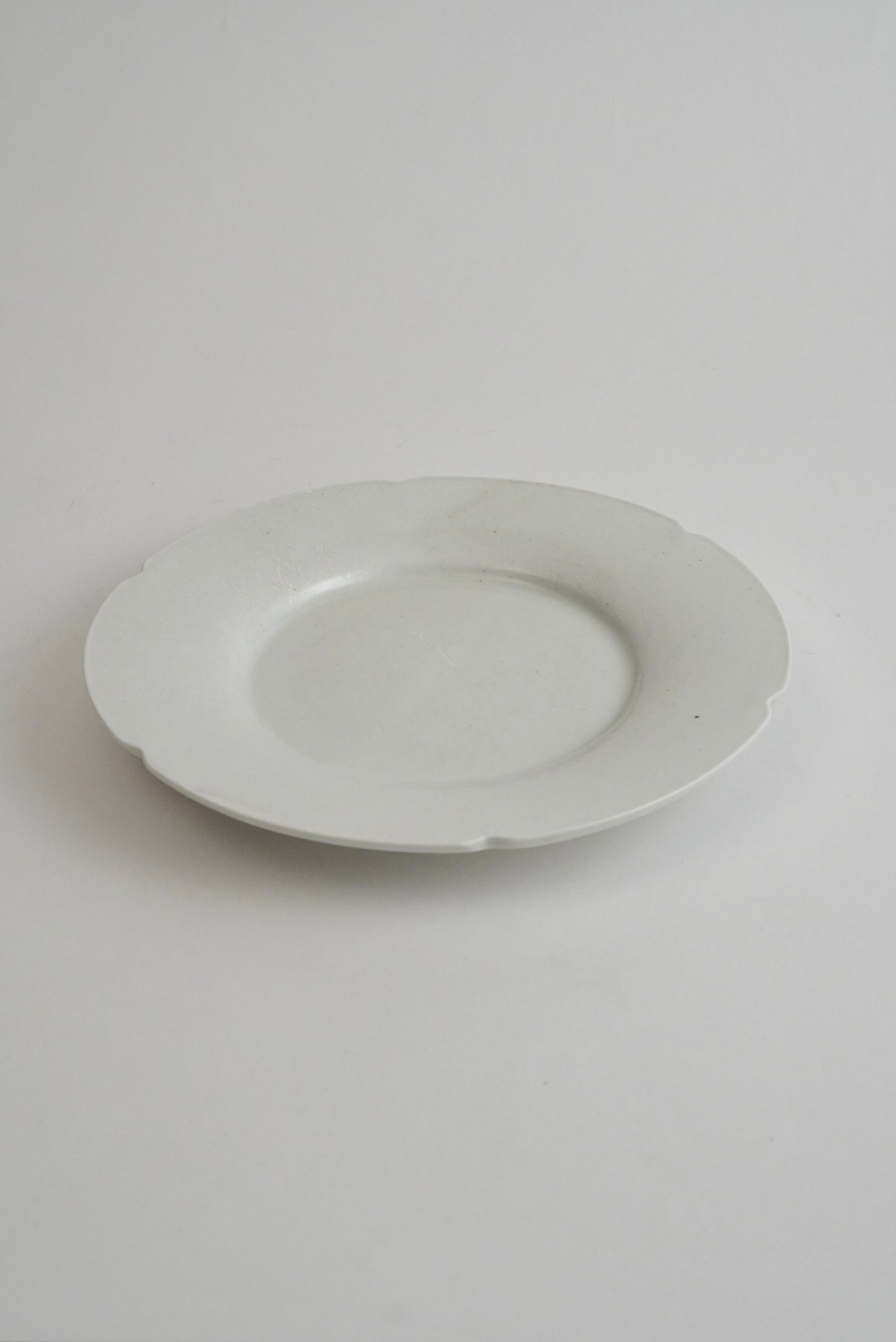 A minimalist white ceramic plate with a scalloped rim, photographed from a top-down angle on a seamless light gray background.