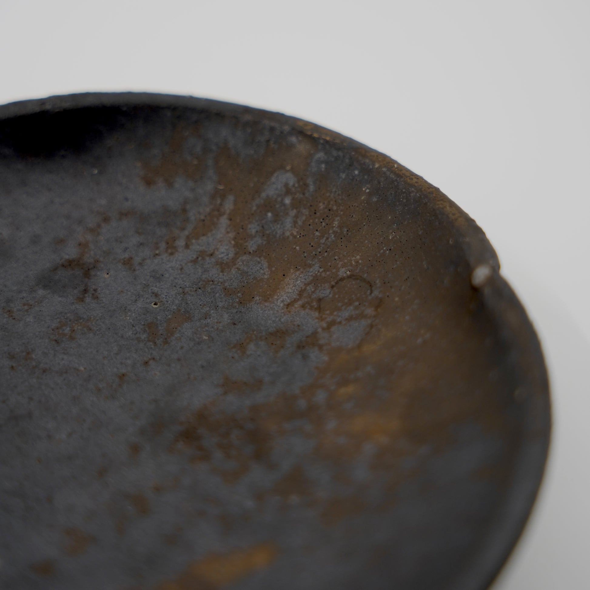 Close-up of a textured dark brown ceramic plate on a light gray background. Handmade ceramic  plate by Japanese artist at Gallery tete Hong Kong.